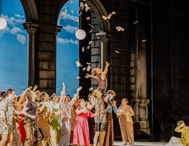 A performer is raised into the air by other performers as they chuck up pieces of paper into the air. Another performer sits on the floor in front of them holding on to their ears. They stand against an archway with street lamps hanging down and the backdrop is painted like a bright sky on a sunny day. (5 of 6)