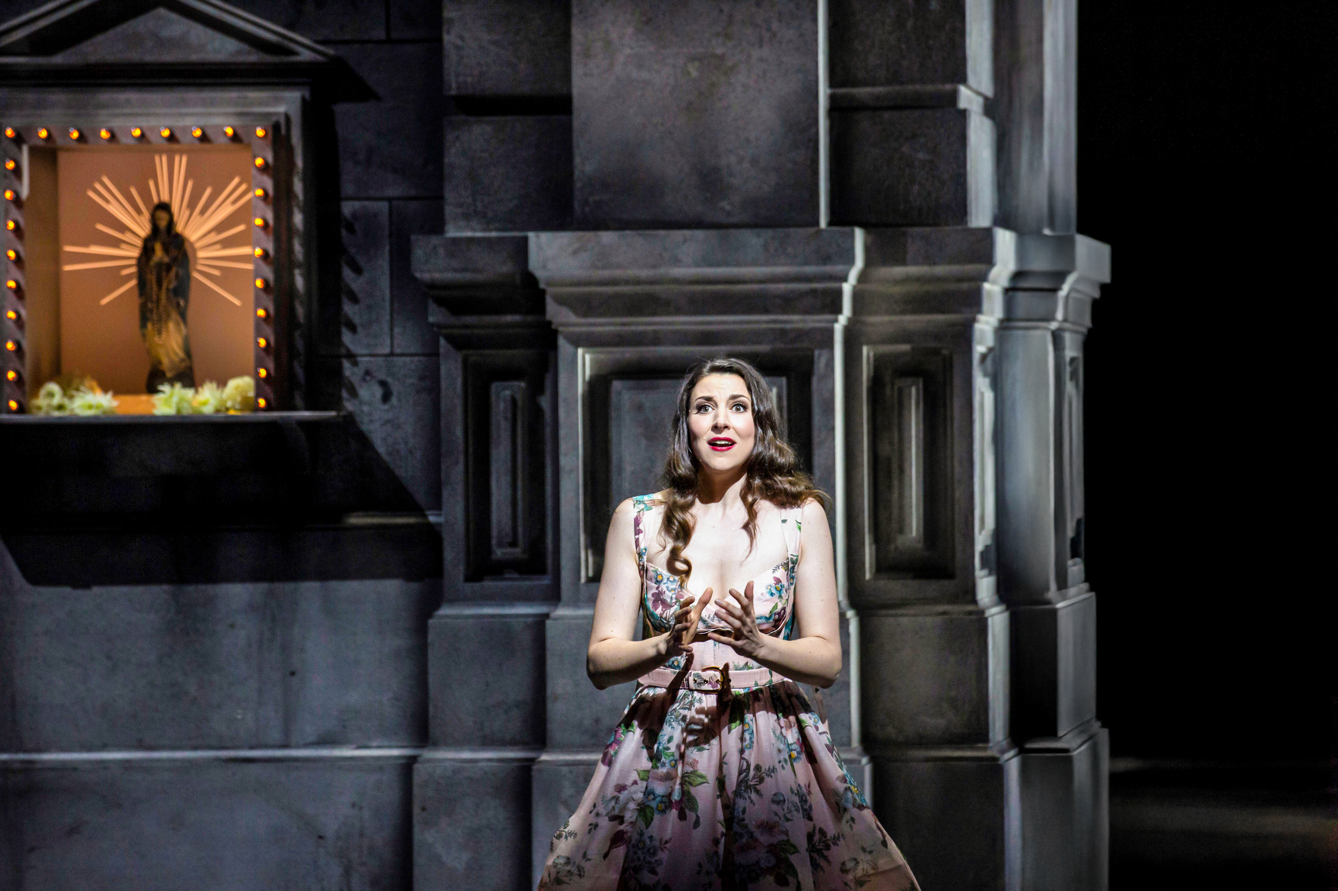 A performer wearing a flowered print dress sits against a church wall. Above her affixed to the wall is a lit sculpture of the Virgin Mary inside a church shrine box. (6 of 6)