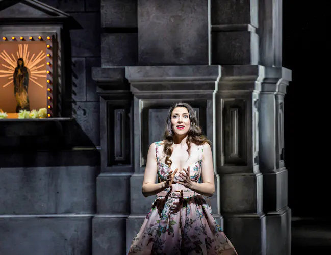 A performer wearing a flowered print dress sits against a church wall. Above her affixed to the wall is a lit sculpture of the Virgin Mary inside a church shrine box. (6 of 6)