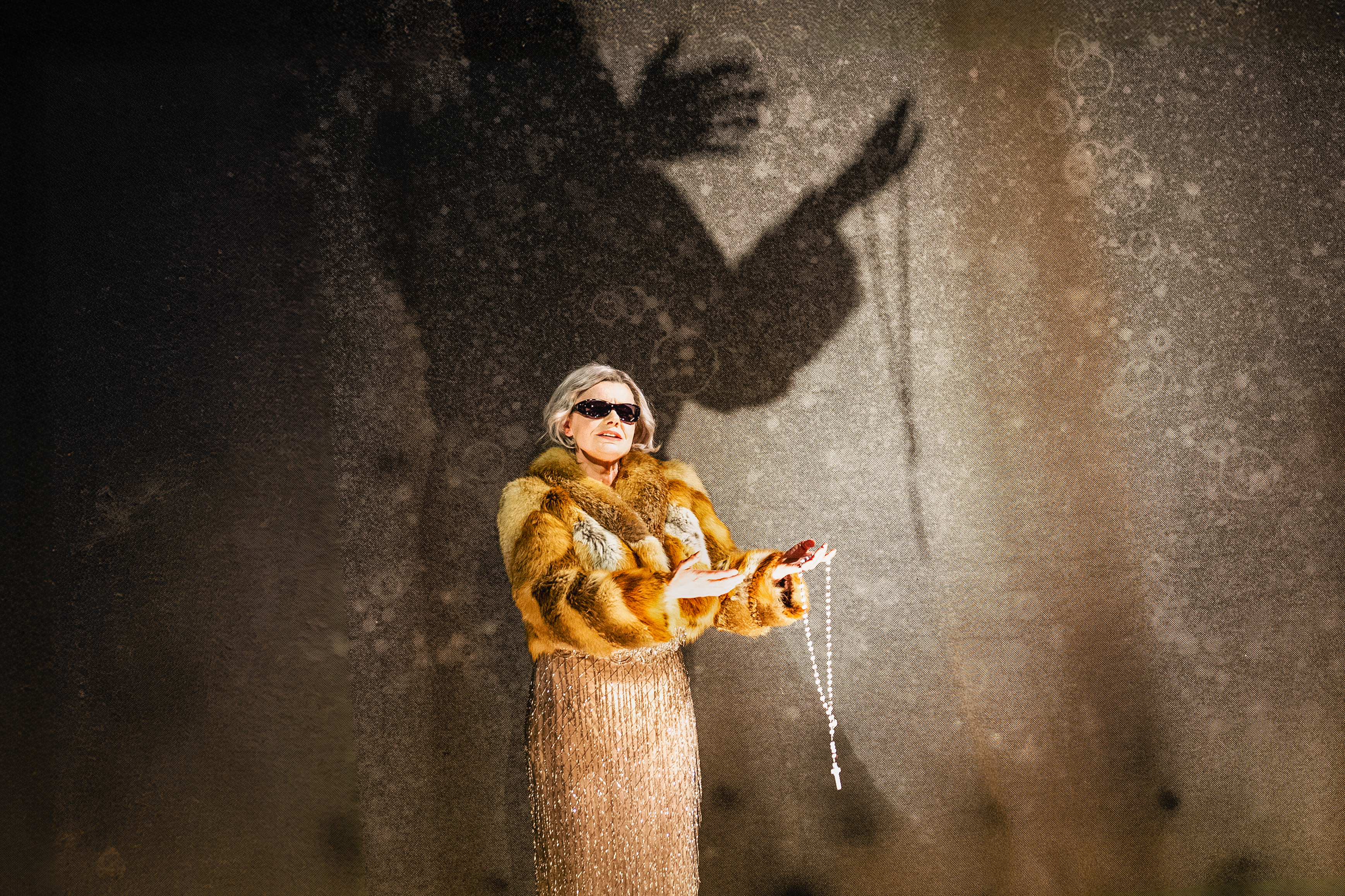 A performer wears a gold sequin tassled dress, a warm-coloured fur coat, black sunglasses and holds a rosary in her left hand. She stands against a stark concrete backdrop with her shadow visibly larger than her. 