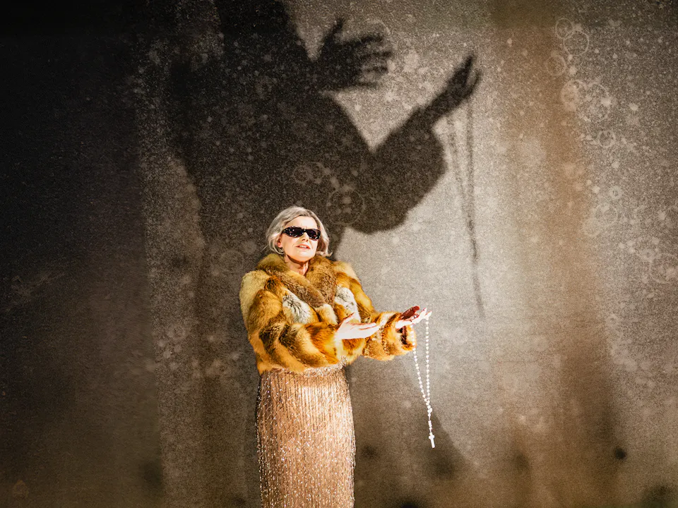 A performer wears a gold sequin tassled dress, a warm-coloured fur coat, black sunglasses and holds a rosary in her left hand. She stands against a stark concrete backdrop with her shadow visibly larger than her.