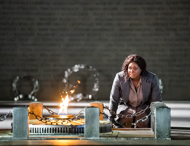 A performer wearing a long grey coat sits on the ground looking at a fire which has been lit upon a memorial. (5 of 12)