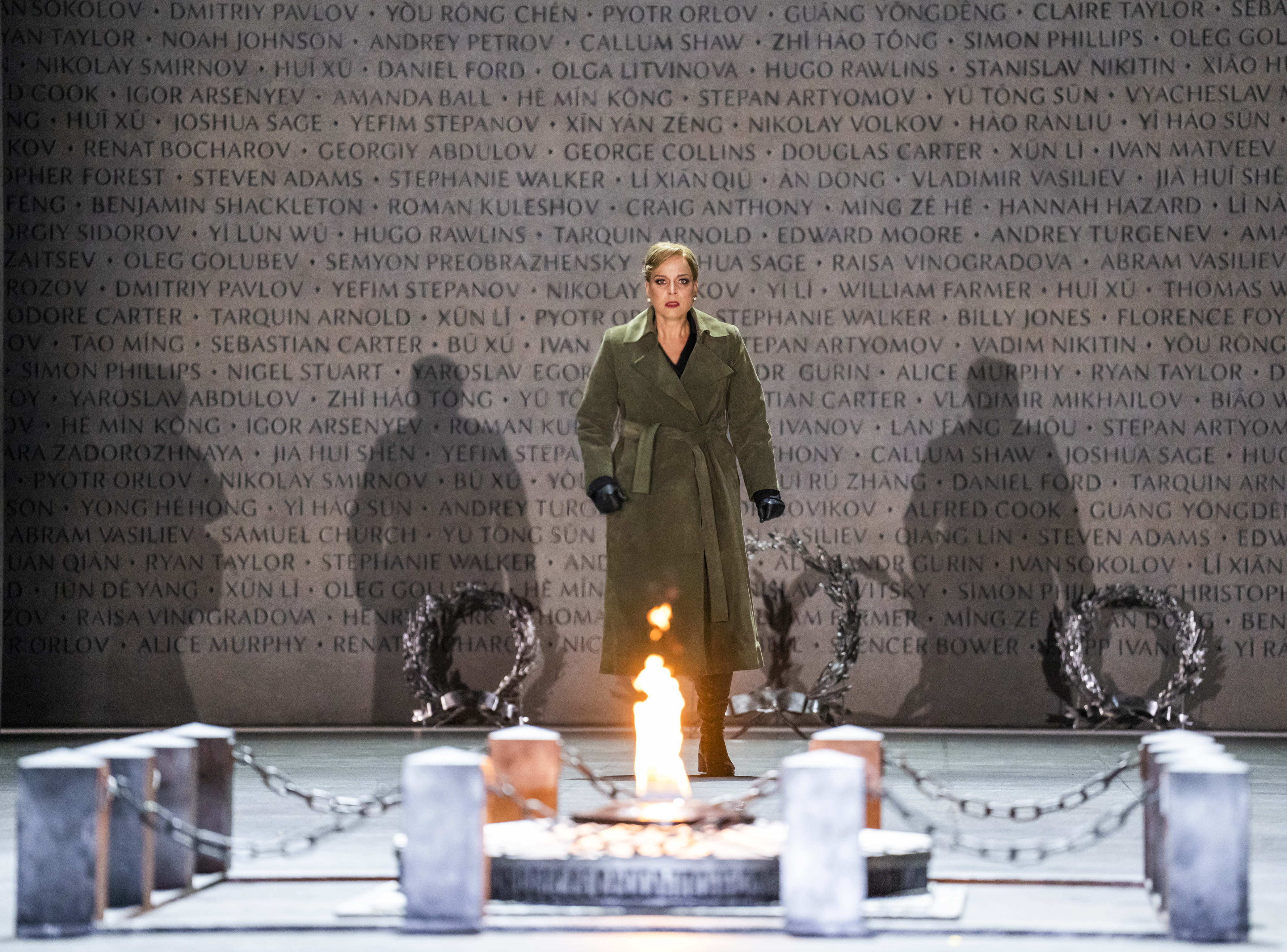 A performer stands looking at a memorial which has a small fire lit upon it. They stand in front of a backdrop of a concrete wall with names engraved into and the shadows of three other people is made visible from the lighting. (2 of 6)