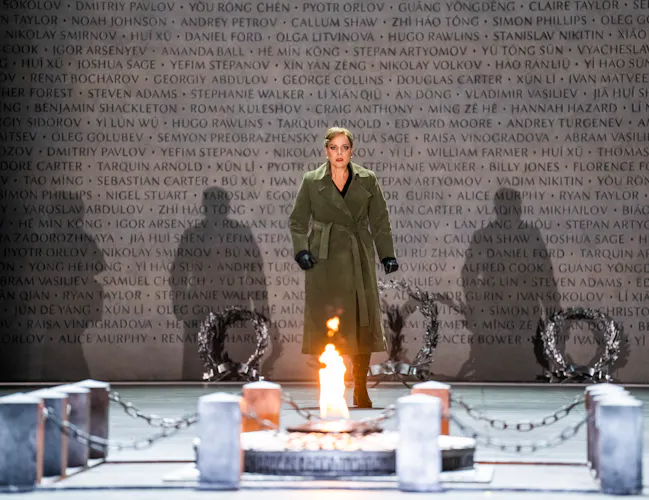 A performer stands looking at a memorial which has a small fire lit upon it. They stand in front of a backdrop of a concrete wall with names engraved into and the shadows of three other people is made visible from the lighting. (2 of 6)