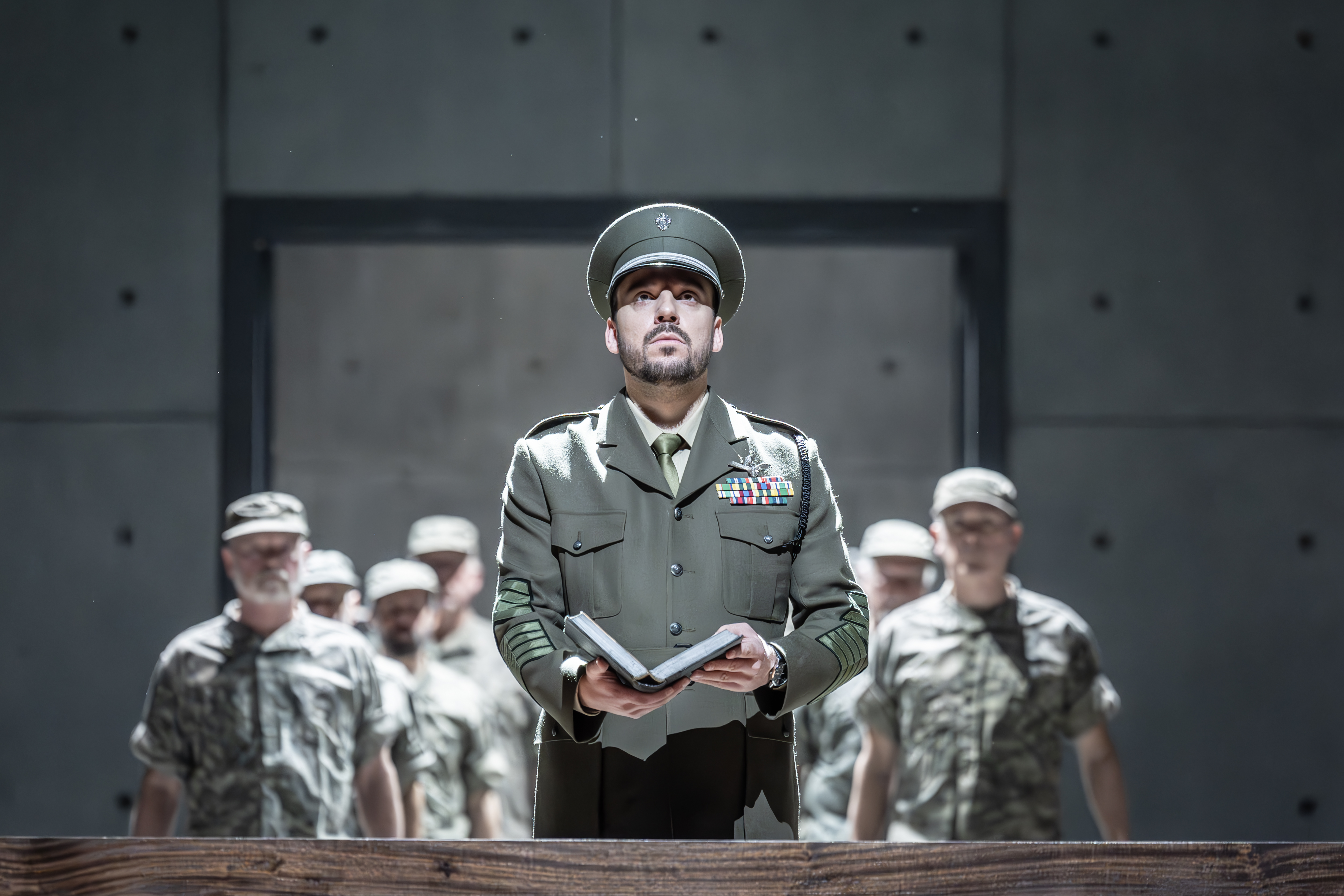 A performer wearing a military uniform holds a book and looks upwards. Behind them several performers wearing army fatigues stand in a line. (3 of 6)