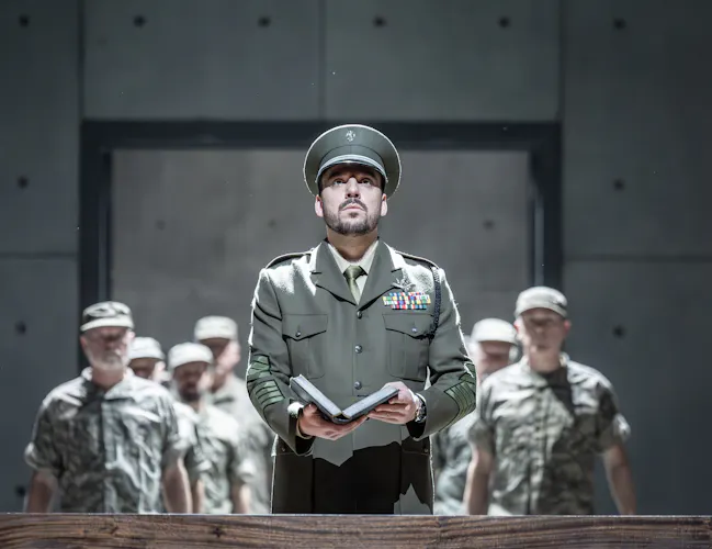 A performer wearing a military uniform holds a book and looks upwards. Behind them several performers wearing army fatigues stand in a line. (3 of 6)