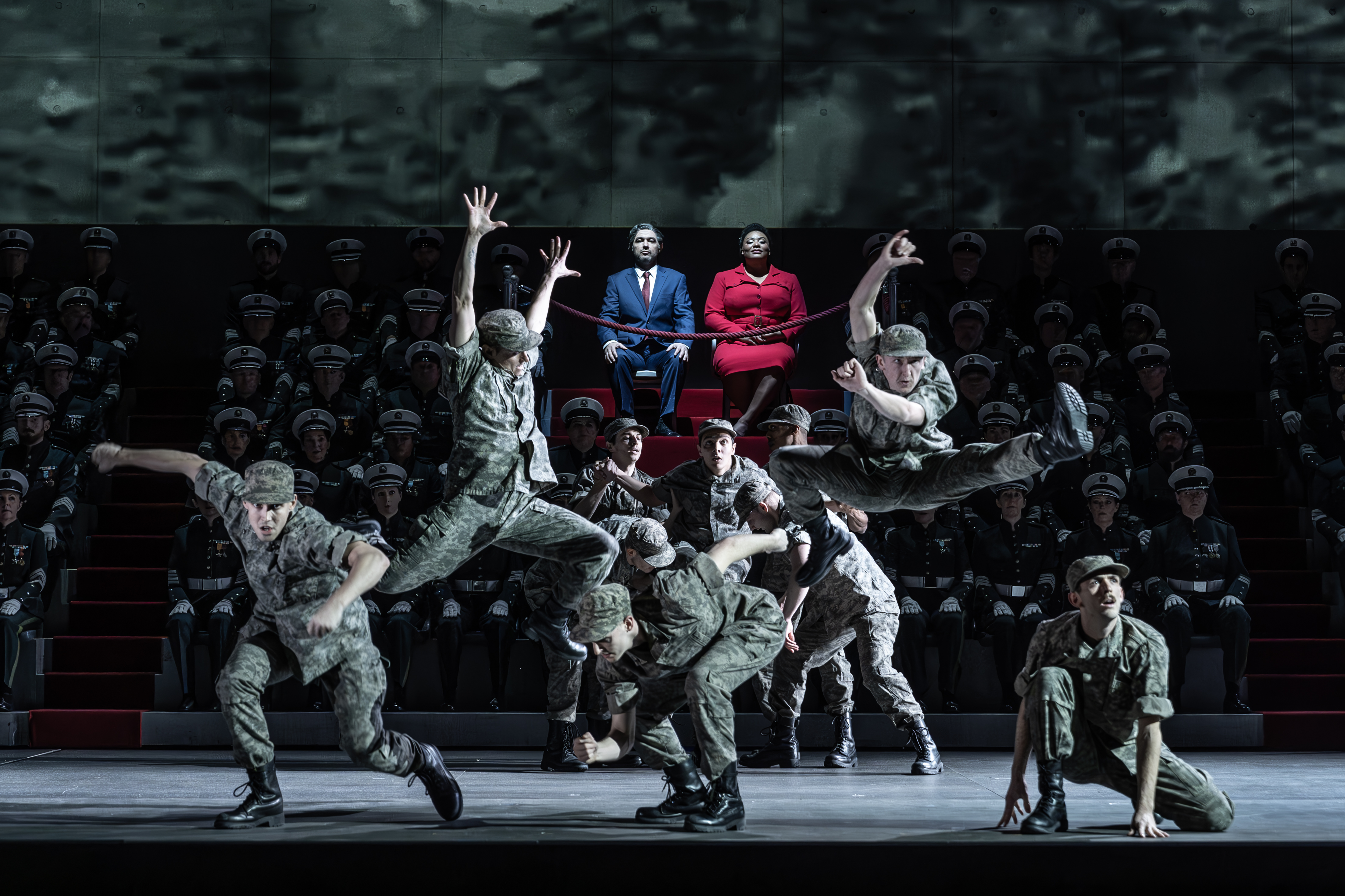 Several performers wearing army fatigues are in movement, with some jumping into the air. They are performing in front of performers sat on arena seating but only two of these performers are visible and lit up - one wears a red suit jacket and skirt and the other wears a blue suit and tie. (4 of 6)