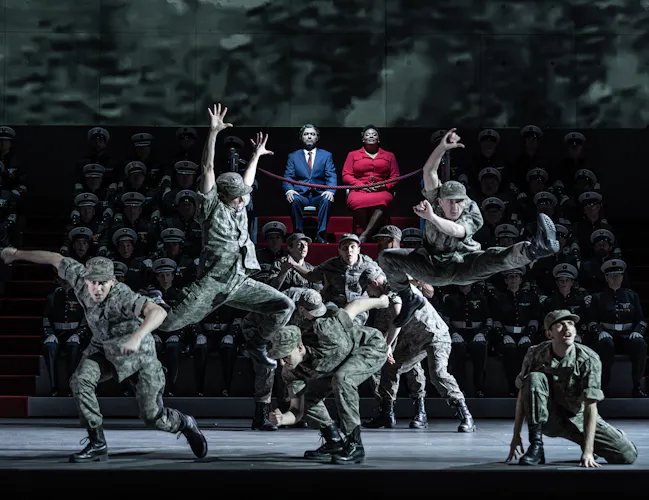 Several performers wearing army fatigues are in movement, with some jumping into the air. They are performing in front of performers sat on arena seating but only two of these performers are visible and lit up - one wears a red suit jacket and skirt and the other wears a blue suit and tie. (4 of 6)