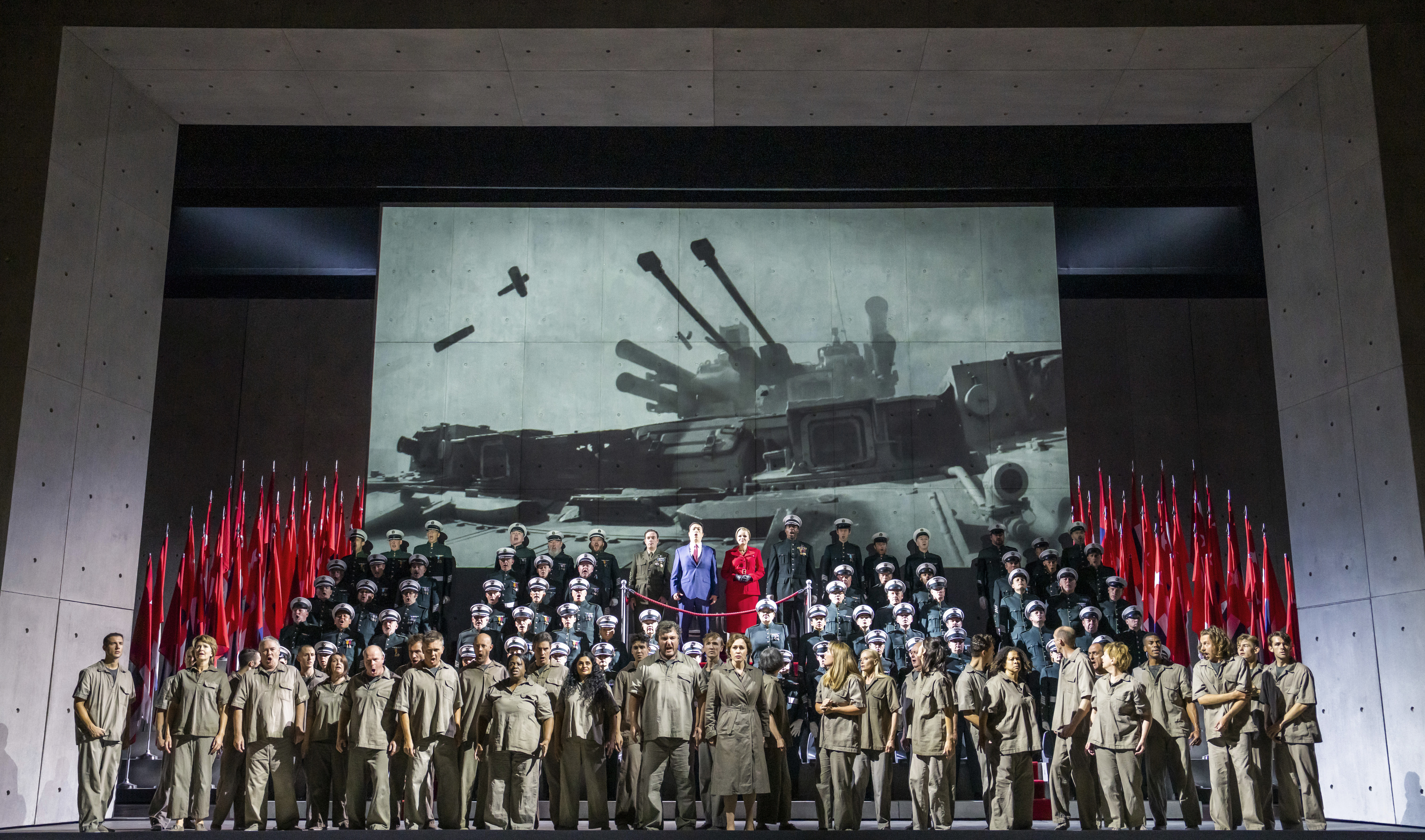 Many performers stand on stage in front of an image of military vehicles projected flanked by several red flats in low mast position. On a platform behind the initial group are a group in military uniforms and in the centre is two performers wearing a red suit jacket and skirt and a blue skirt and tie. (5 of 6)