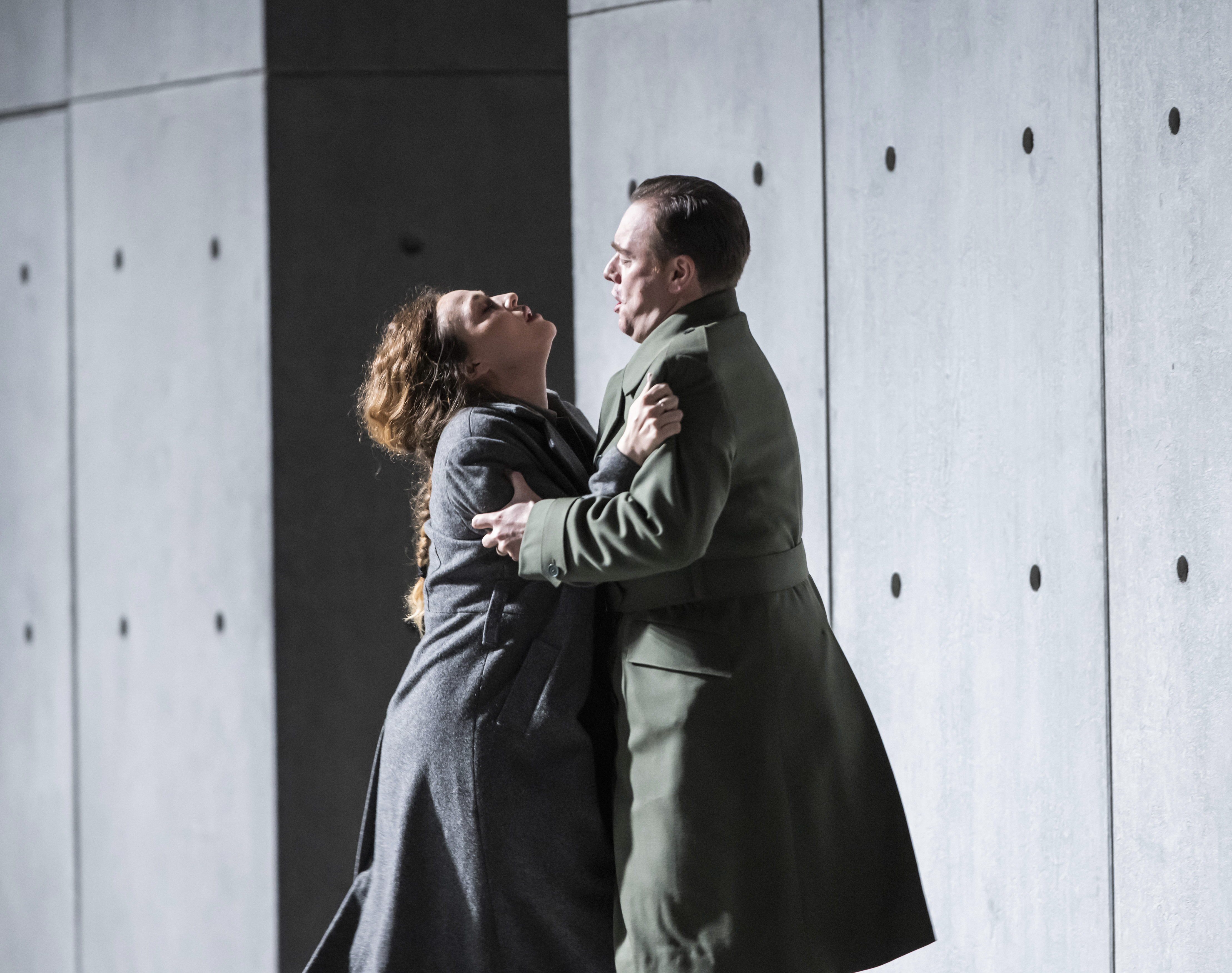 Two performers hold each other passionately on stage in front of a stark concrete wall. (6 of 6)