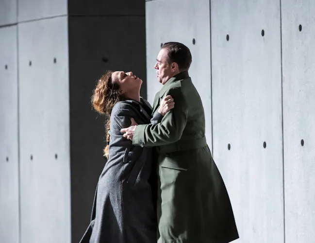 Two performers hold each other passionately on stage in front of a stark concrete wall. (6 of 6)