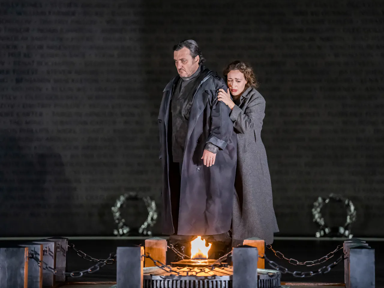 A performer wearing a long jacket is embraced from behind by another. They both stare down at a small fire atop a memorial stone.