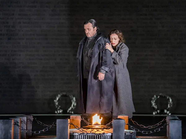 A performer wearing a long jacket is embraced from behind by another. They both stare down at a small fire atop a memorial stone.