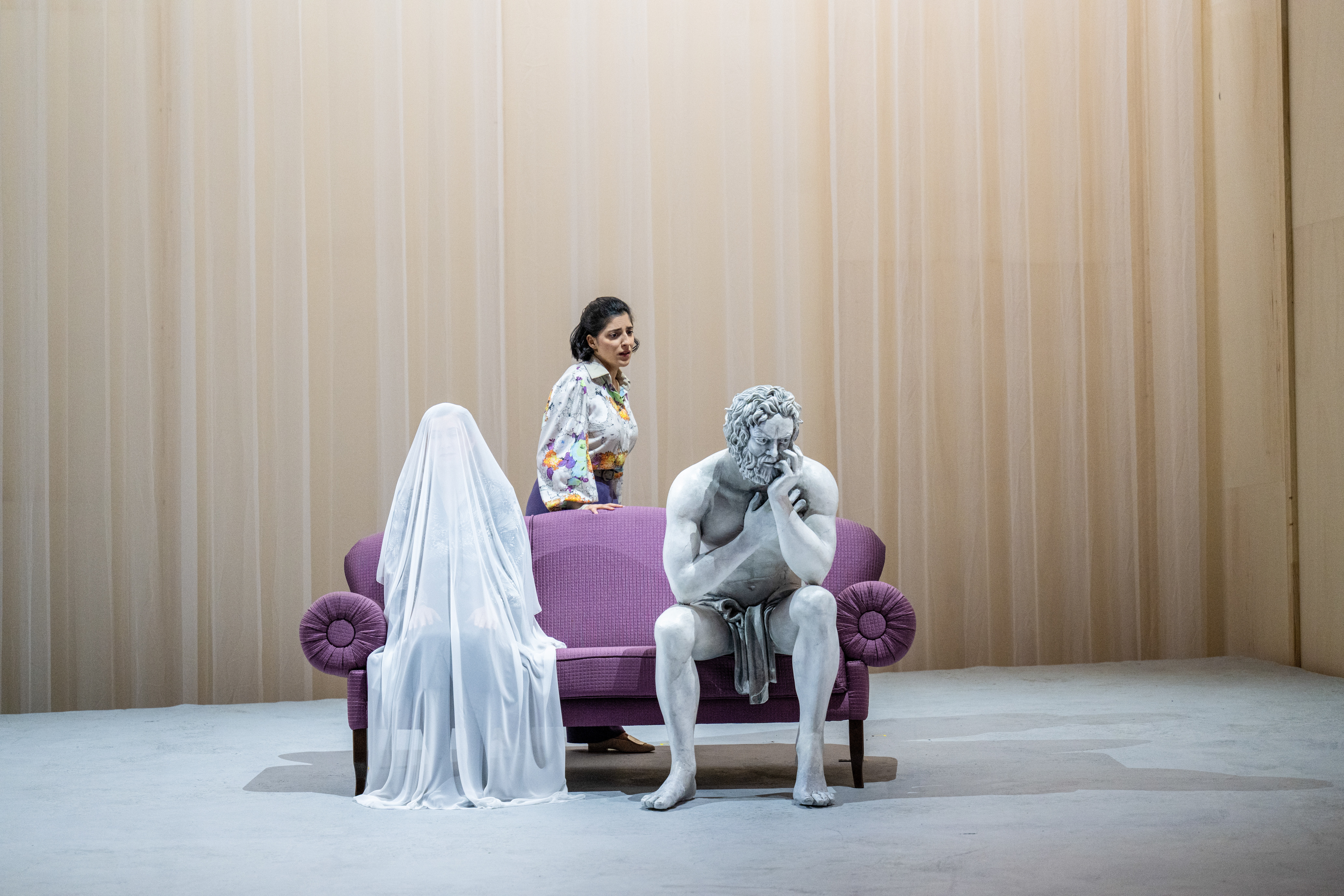 A performer whose makeup and costume is designed to make them look like a statue sits on a purple sofa on stage with a white curtain as a backdrop. Next to them sits a performer beneath a full length white veil, with their body only just visible. A performer wearing a patterned shirt stands behind the sofa. (7 of 12)