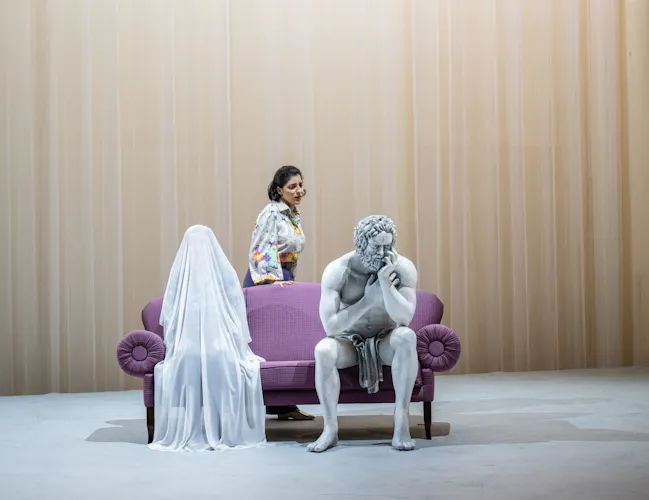 A performer whose makeup and costume is designed to make them look like a statue sits on a purple sofa on stage with a white curtain as a backdrop. Next to them sits a performer beneath a full length white veil, with their body only just visible. A performer wearing a patterned shirt stands behind the sofa. (7 of 12)
