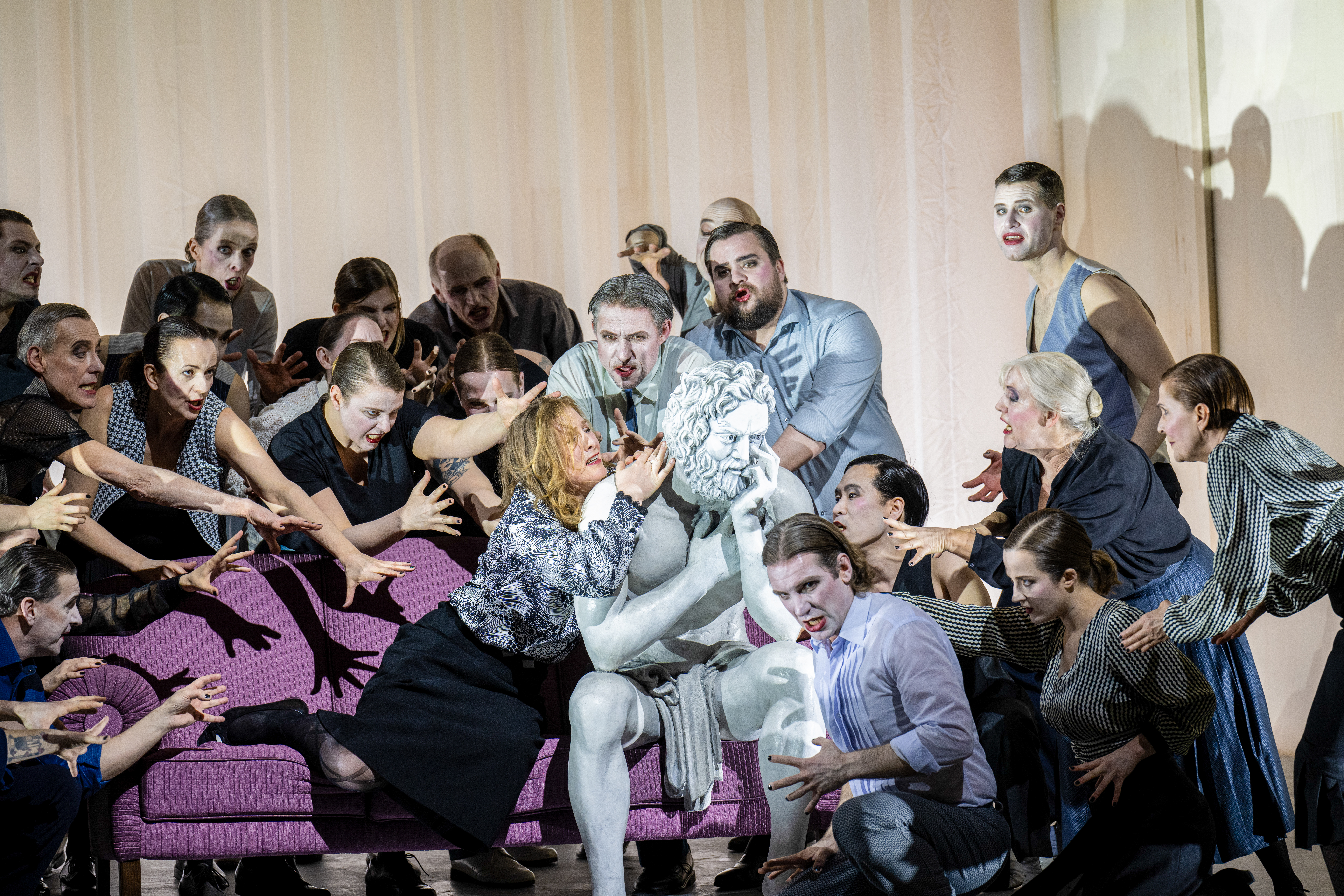 A performer whose makeup and costume is designed to make them look like a statue sits on a purple sofa on stage with a white curtain as a backdrop. They are surrounded by a group of people who reach out to touch them. (1 of 5)