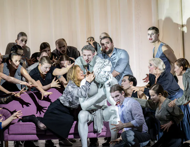 A performer whose makeup and costume is designed to make them look like a statue sits on a purple sofa on stage with a white curtain as a backdrop. They are surrounded by a group of people who reach out to touch them. (1 of 5)