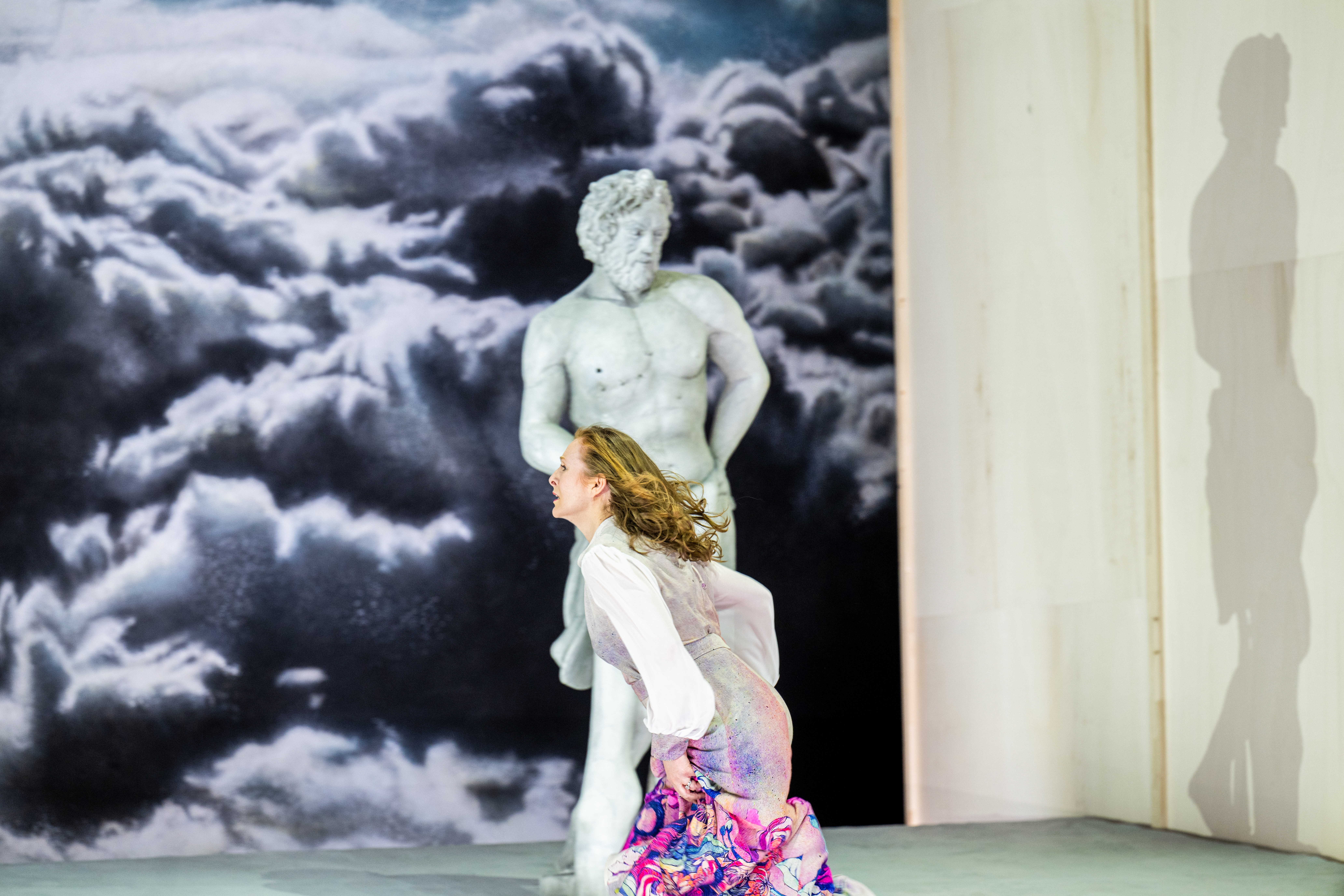 A performer whose makeup and costume is designed to make them look like a statue stands up against a backdrop of a painted cloudy sky, their shadow visible next to them. A person wearing a pink patterned dress runs past them. (2 of 5)