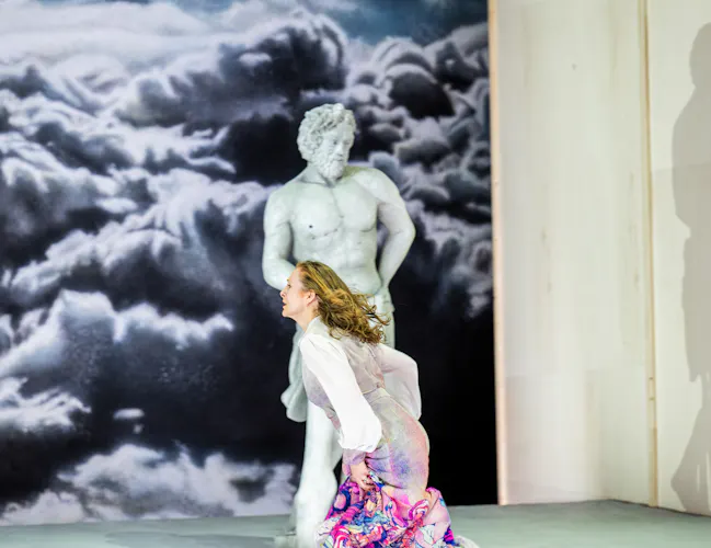 A performer whose makeup and costume is designed to make them look like a statue stands up against a backdrop of a painted cloudy sky, their shadow visible next to them. A person wearing a pink patterned dress runs past them. (2 of 5)