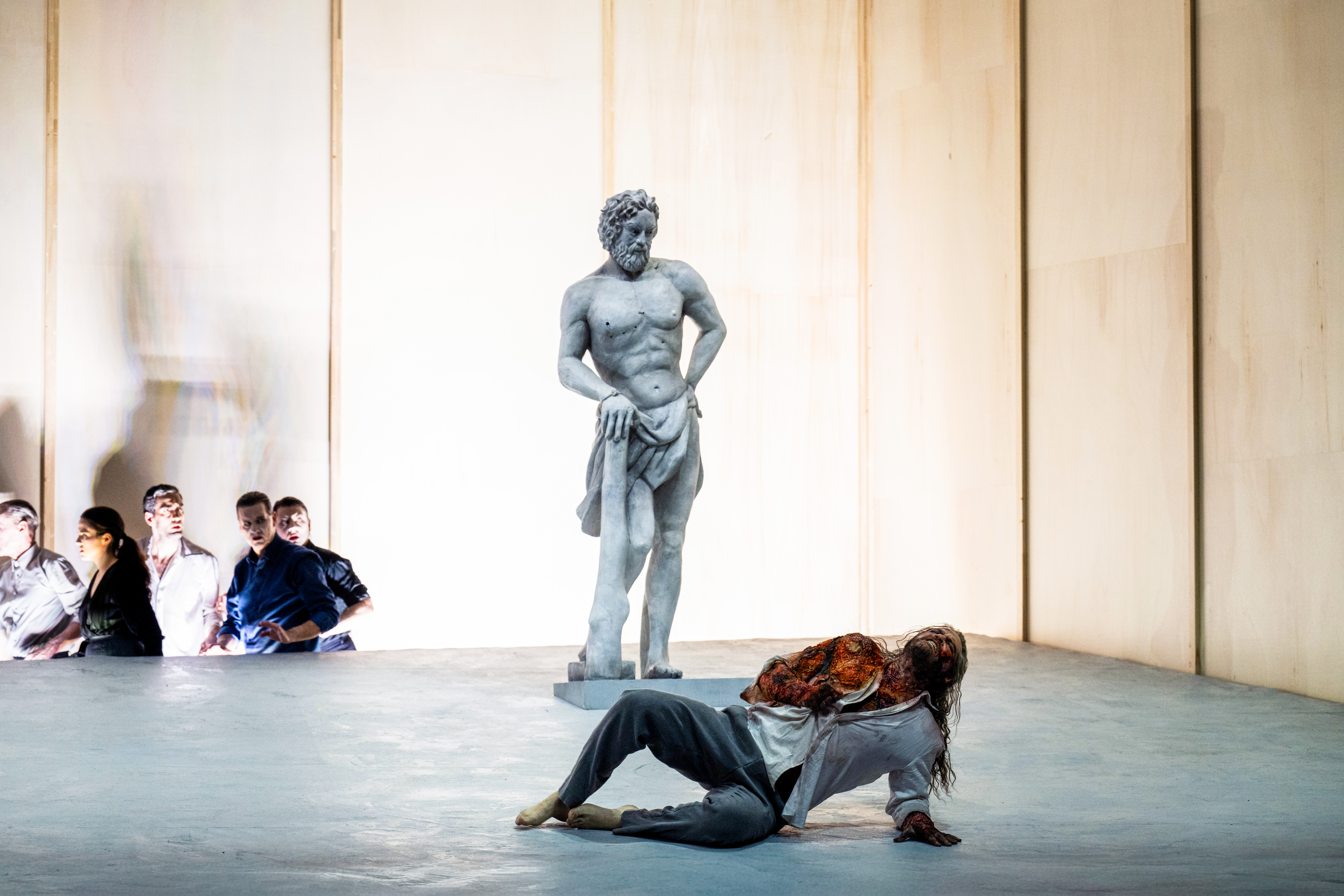 A performer whose makeup and costume is designed to make them look like a statue stands up against a stark backdrop. A person wearing a white patterned shirt lies on the floor in front of them, appearing to writhe in pain. (3 of 5)