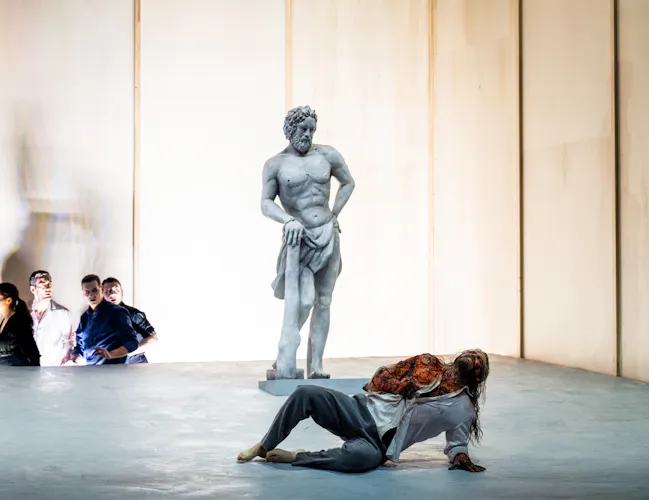 A performer whose makeup and costume is designed to make them look like a statue stands up against a stark backdrop. A person wearing a white patterned shirt lies on the floor in front of them, appearing to writhe in pain. (3 of 5)