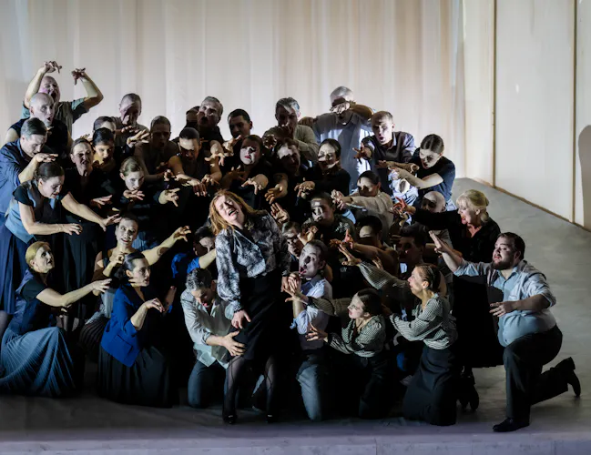 A group of performers stand one performer wearing a check shirt on stage. They are all holding their arms up towards them. (4 of 5)