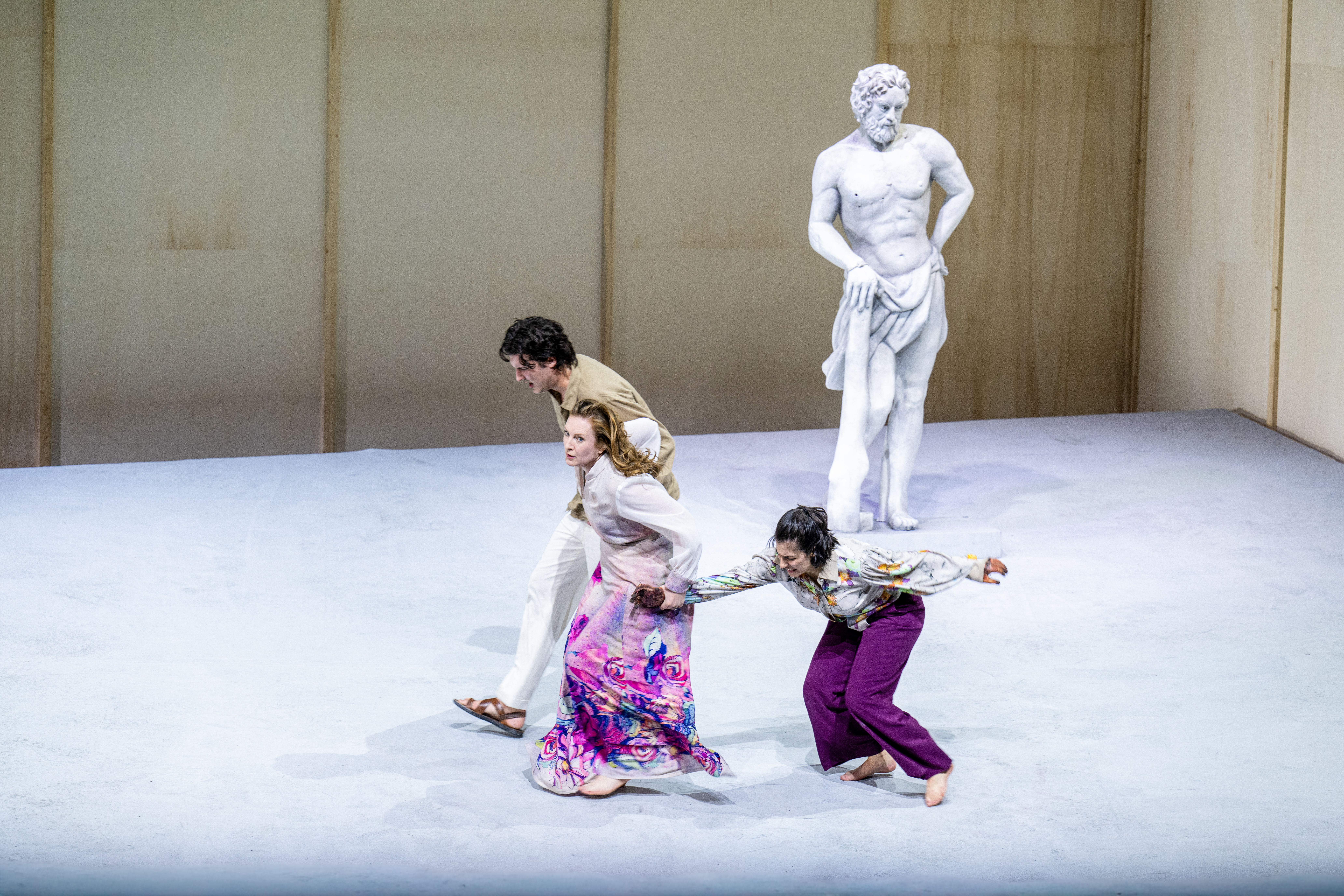 A performer whose makeup and costume is designed to make them look like a statue stands on stage like a statue. Three performers wearing bright clothes run past. (5 of 5)