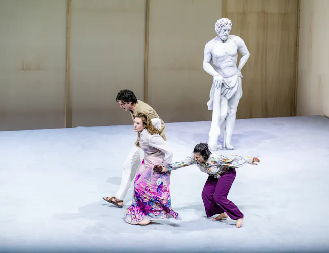 A performer whose makeup and costume is designed to make them look like a statue stands on stage like a statue. Three performers wearing bright clothes run past. (5 of 5)