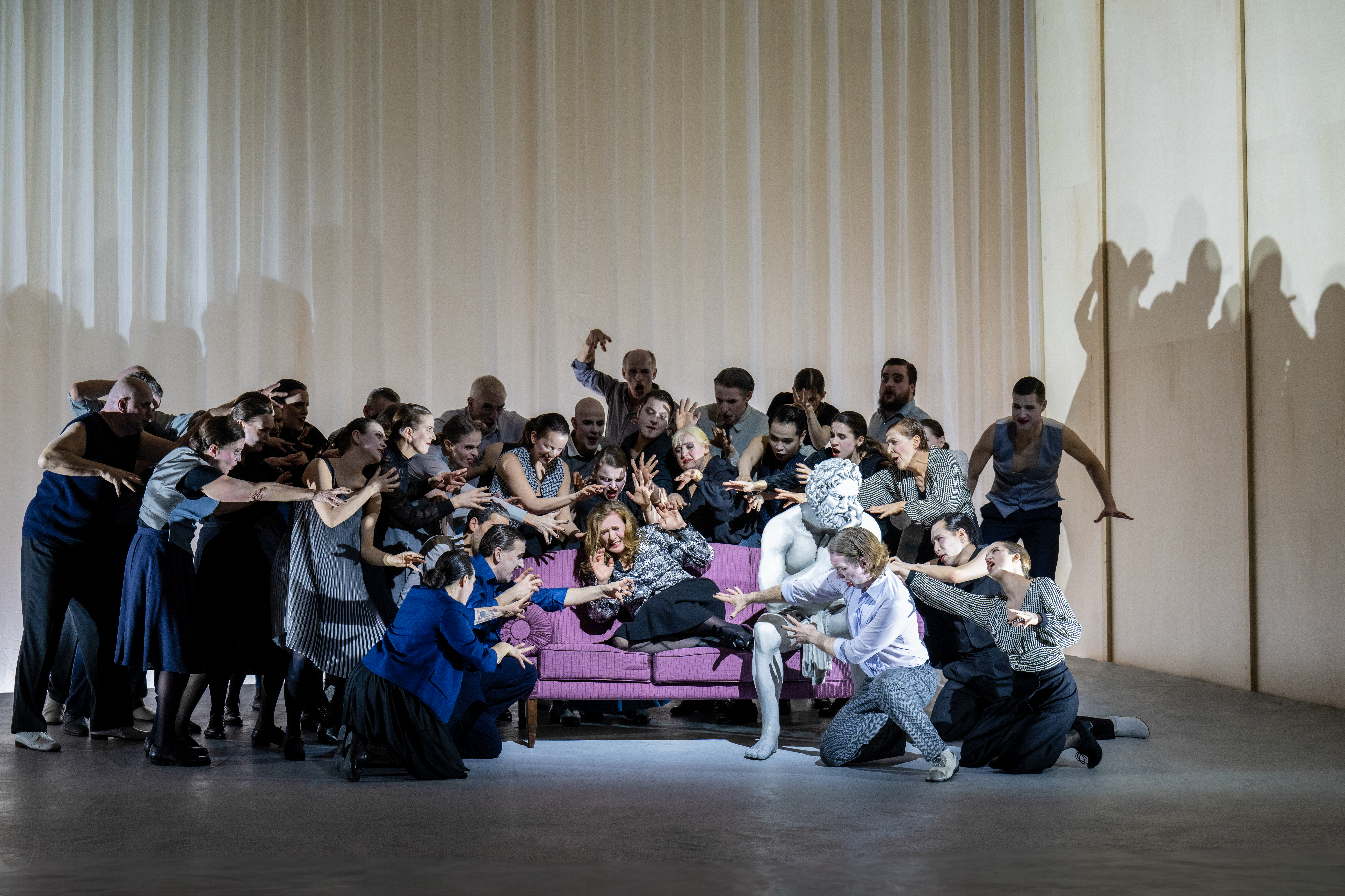 A performer whose makeup and costume is designed to make them look like a statue sits on a purple sofa on stage. Next to them sits another performer with long hair wearing a check shirt. Around them are a group of people reaching out towards the performer in the check shirt, as the statue seems to be ignored. 