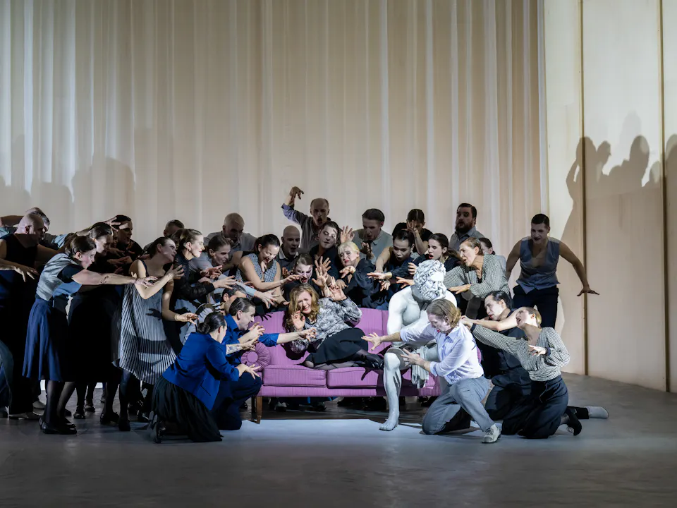 A performer whose makeup and costume is designed to make them look like a statue sits on a purple sofa on stage. Next to them sits another performer with long hair wearing a check shirt. Around them are a group of people reaching out towards the performer in the check shirt, as the statue seems to be ignored.