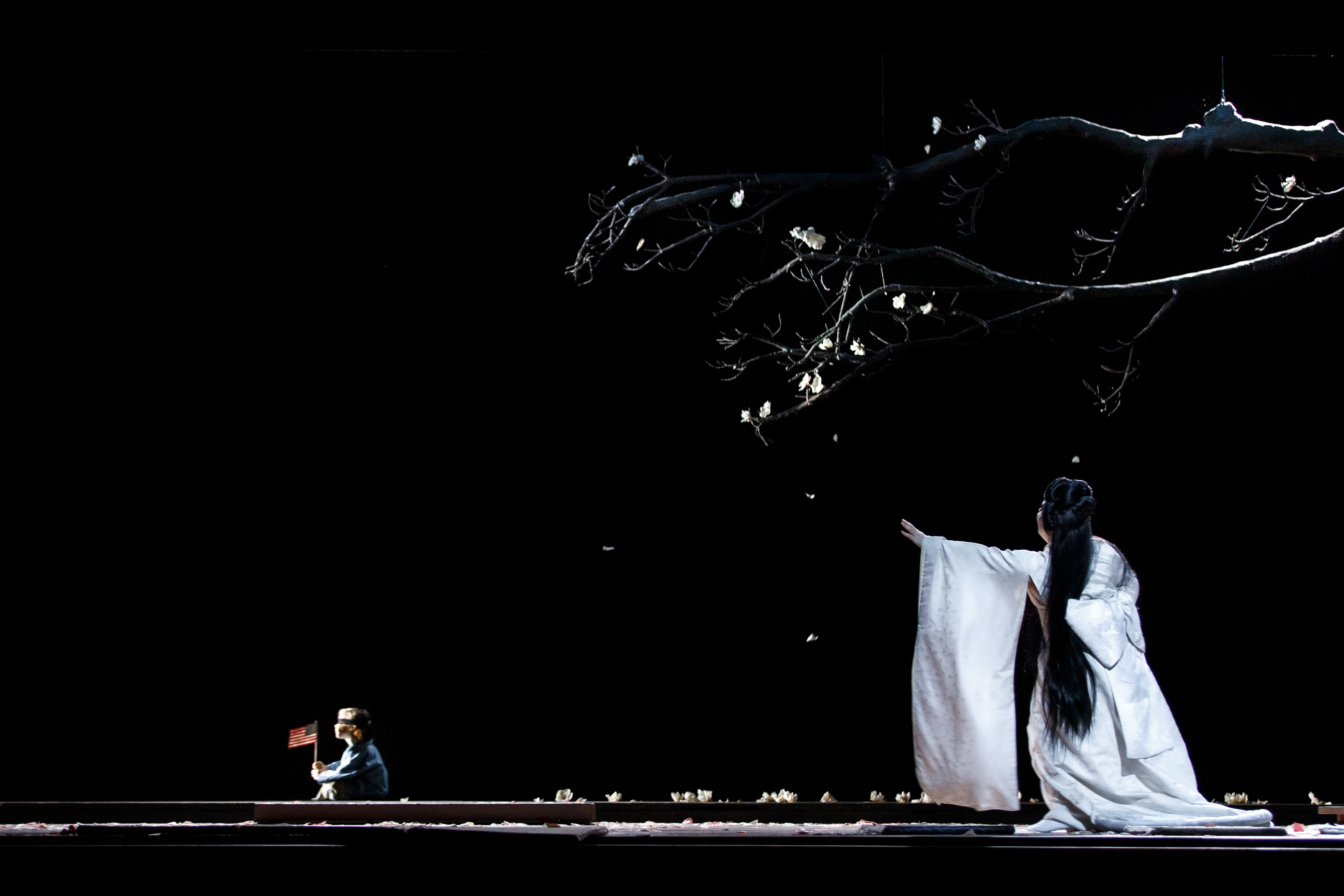 A performer wearing a flowing white Kimono with long brown hair stands on a dark stage beneath a tree, with their arms raised forwards. At the other side of the stage sits a small child. They are performing in The Royal Opera's production of Madama Butterfly. (1 of 6)