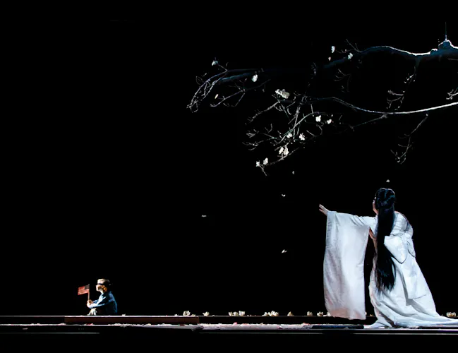A performer wearing a flowing white Kimono with long brown hair stands on a dark stage beneath a tree, with their arms raised forwards. At the other side of the stage sits a small child. They are performing in The Royal Opera's production of Madama Butterfly. (1 of 6)
