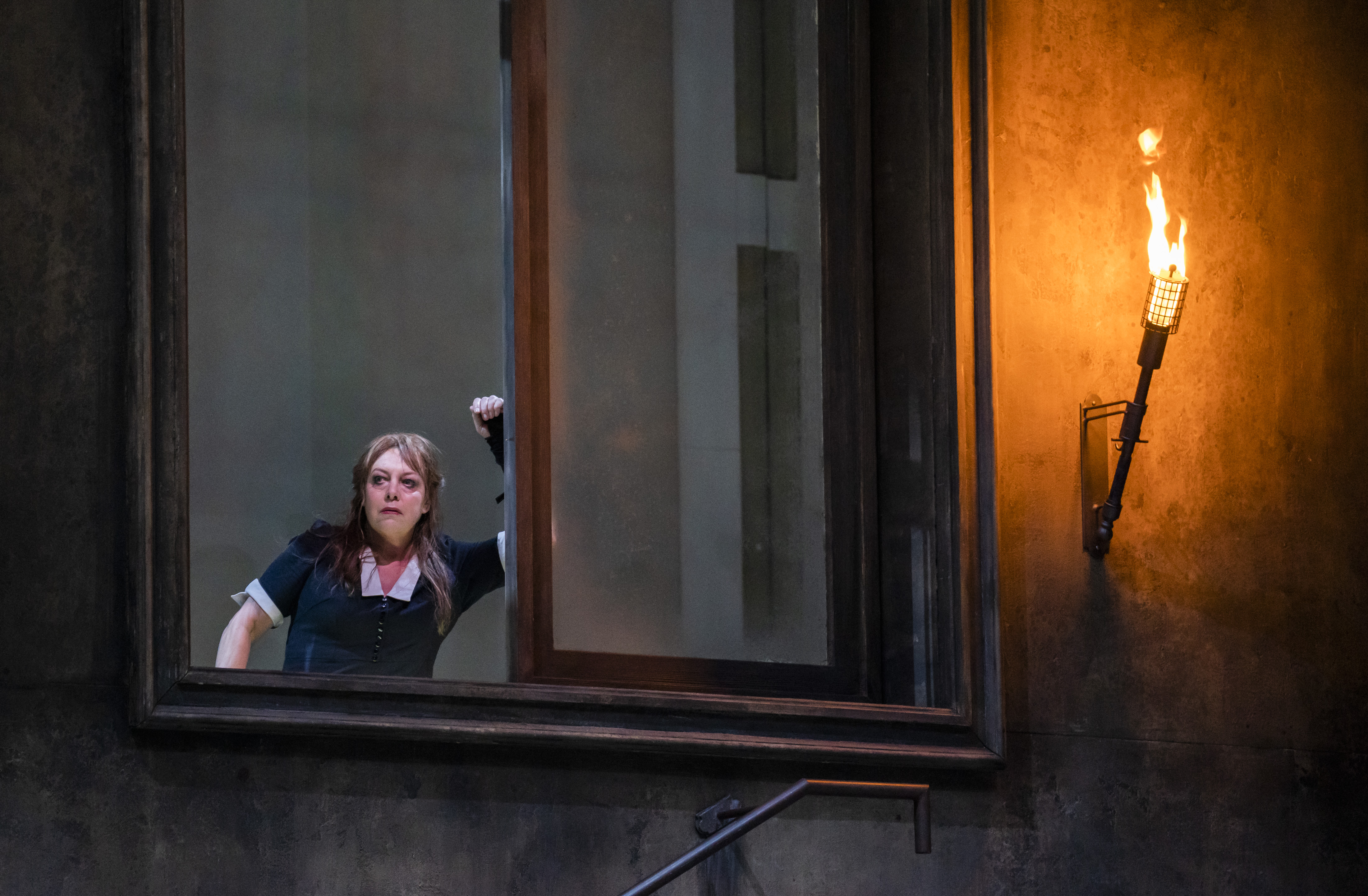 A performer wearing a black and white maids uniform looks out of a window. Next to the window, a lantern is lit up with fire. (3 of 5)