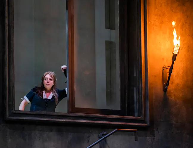 A performer wearing a black and white maids uniform looks out of a window. Next to the window, a lantern is lit up with fire. (3 of 5)