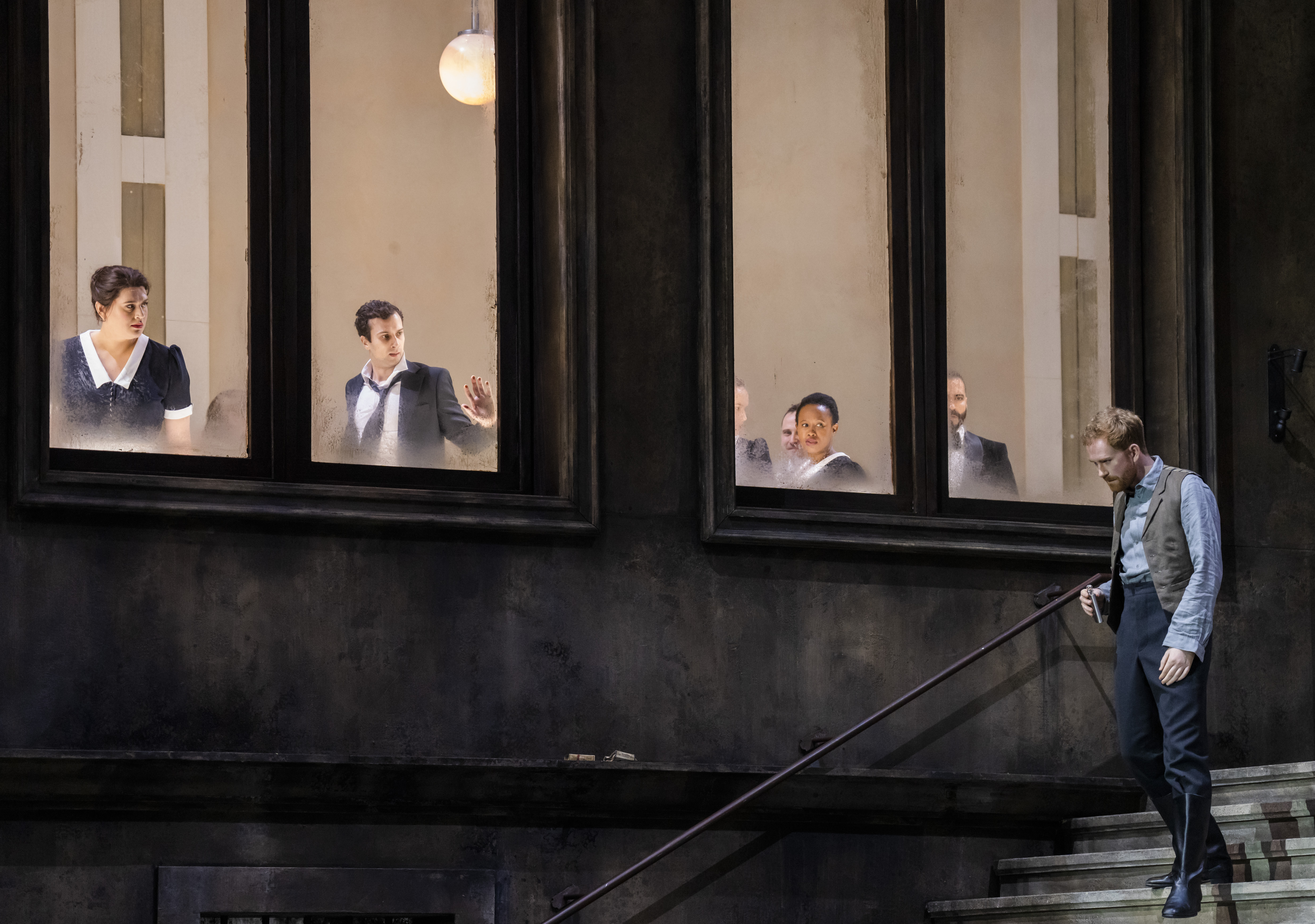 A performer stands on some stairs on a stage, looking down wistfully. Above them, several performers look out of a well-lit window down at him. 