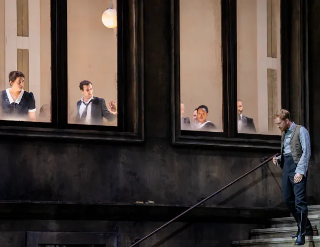 A performer stands on some stairs on a stage, looking down wistfully. Above them, several performers look out of a well-lit window down at him.