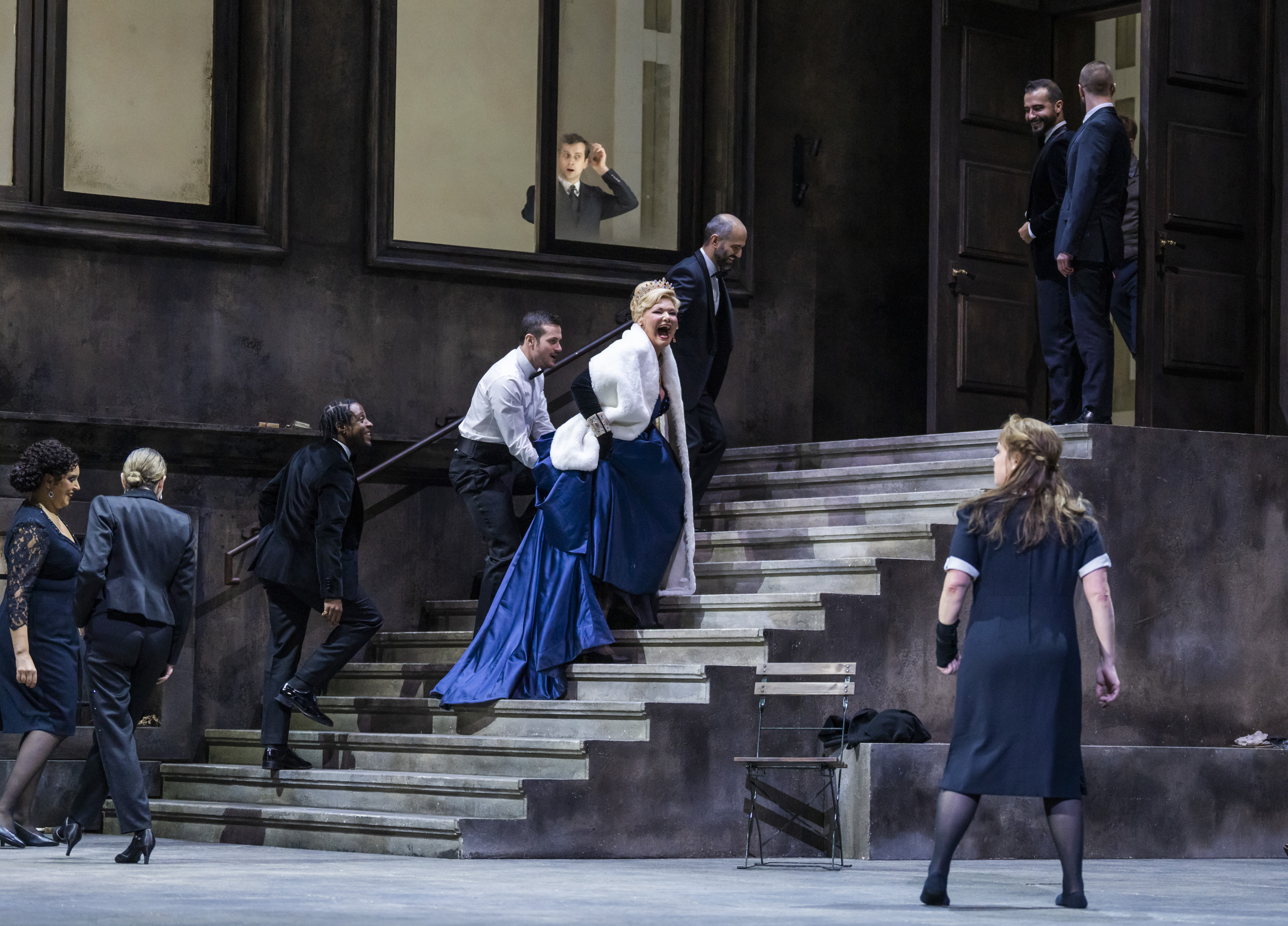 A performer wearing a navy blue ballgown and white fur jacket walks up some concrete stairs on a stage appearing to scream at another performer wearing a black and white maids uniform. They are flanked by performers wearing police uniforms. At the window above, a person looks down at the scene unfolding. 