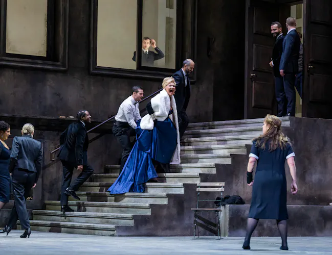 A performer wearing a navy blue ballgown and white fur jacket walks up some concrete stairs on a stage appearing to scream at another performer wearing a black and white maids uniform. They are flanked by performers wearing police uniforms. At the window above, a person looks down at the scene unfolding. (2 of 5)