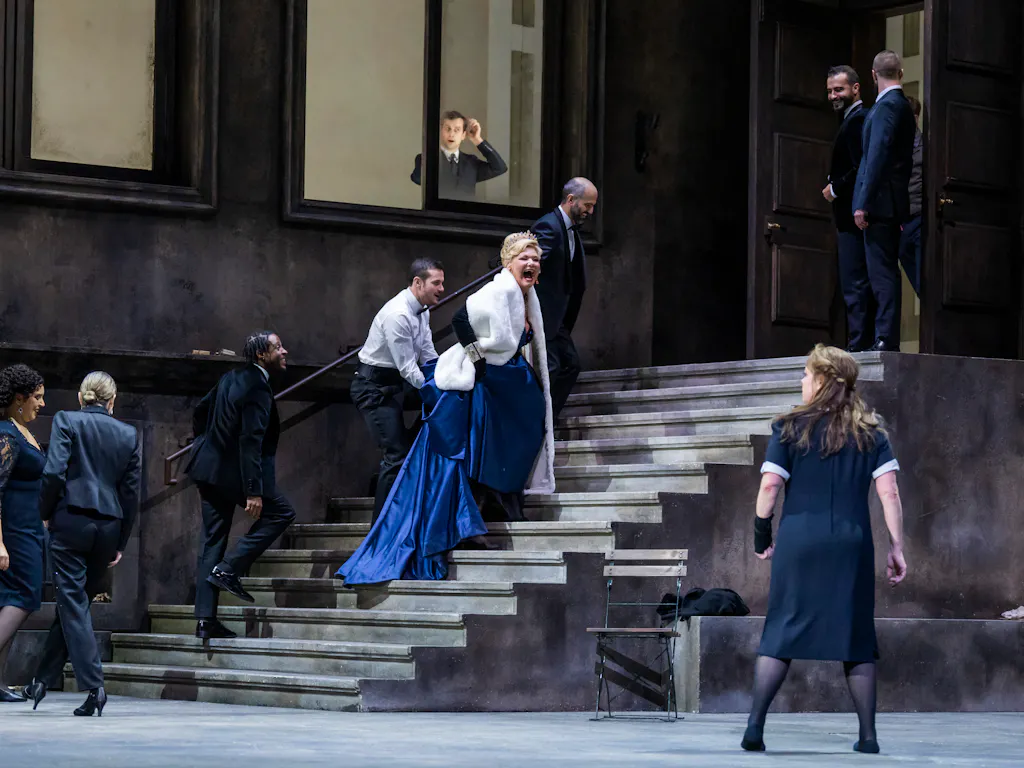 A performer wearing a navy blue ballgown and white fur jacket walks up some concrete stairs on a stage appearing to scream at another performer wearing a black and white maids uniform. They are flanked by performers wearing police uniforms. At the window above, a person looks down at the scene unfolding.