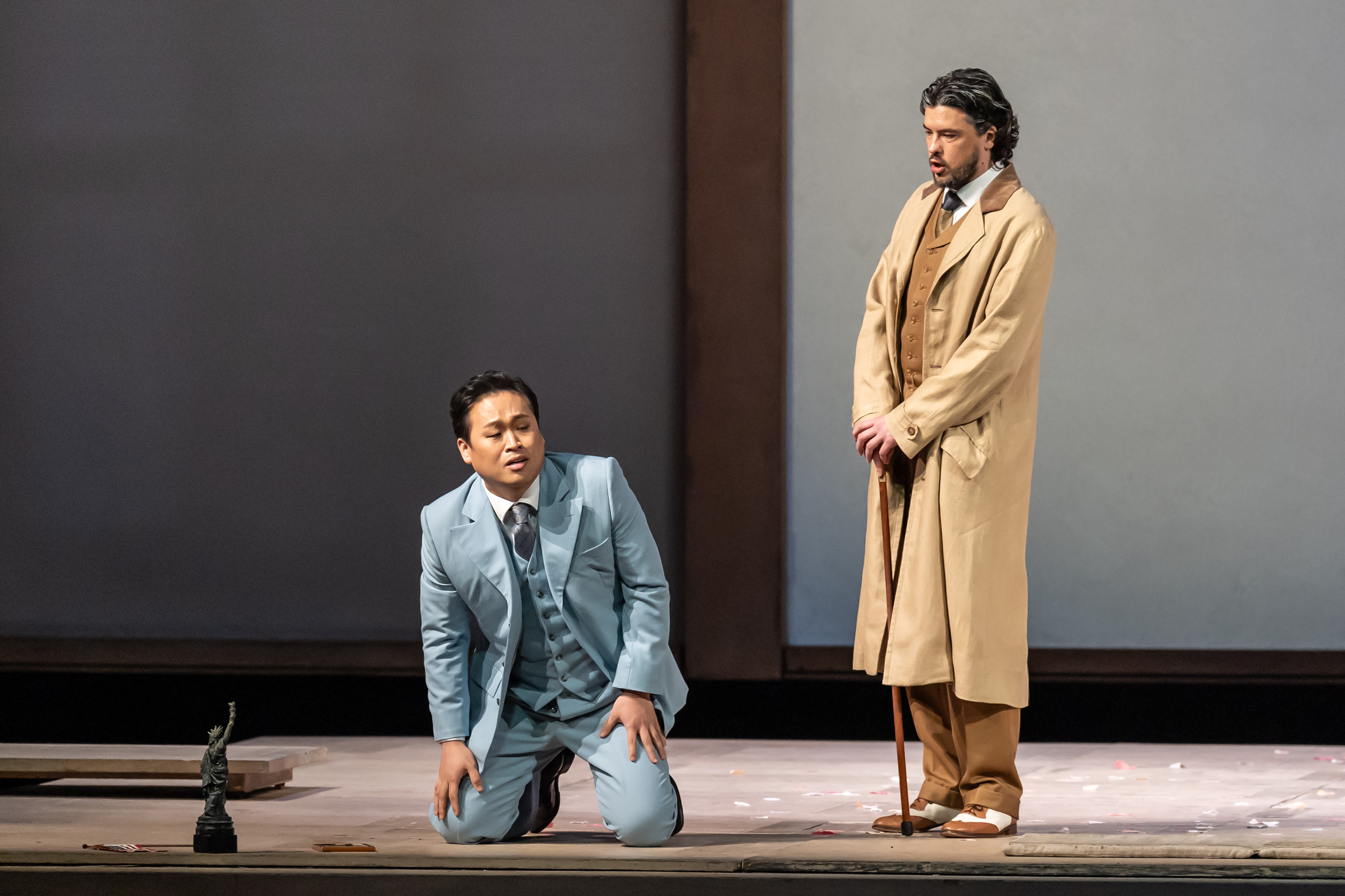 A performer wearing a blue suit sits on their knees on stage. Another wearing a brown suit with a beige dress jacket leans on a cane next to them. They are performing in The Royal Opera's production of Madama Butterfly. (2 of 6)