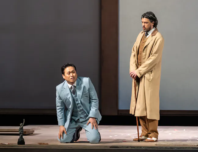 A performer wearing a blue suit sits on their knees on stage. Another wearing a brown suit with a beige dress jacket leans on a cane next to them. They are performing in The Royal Opera's production of Madama Butterfly. (2 of 6)