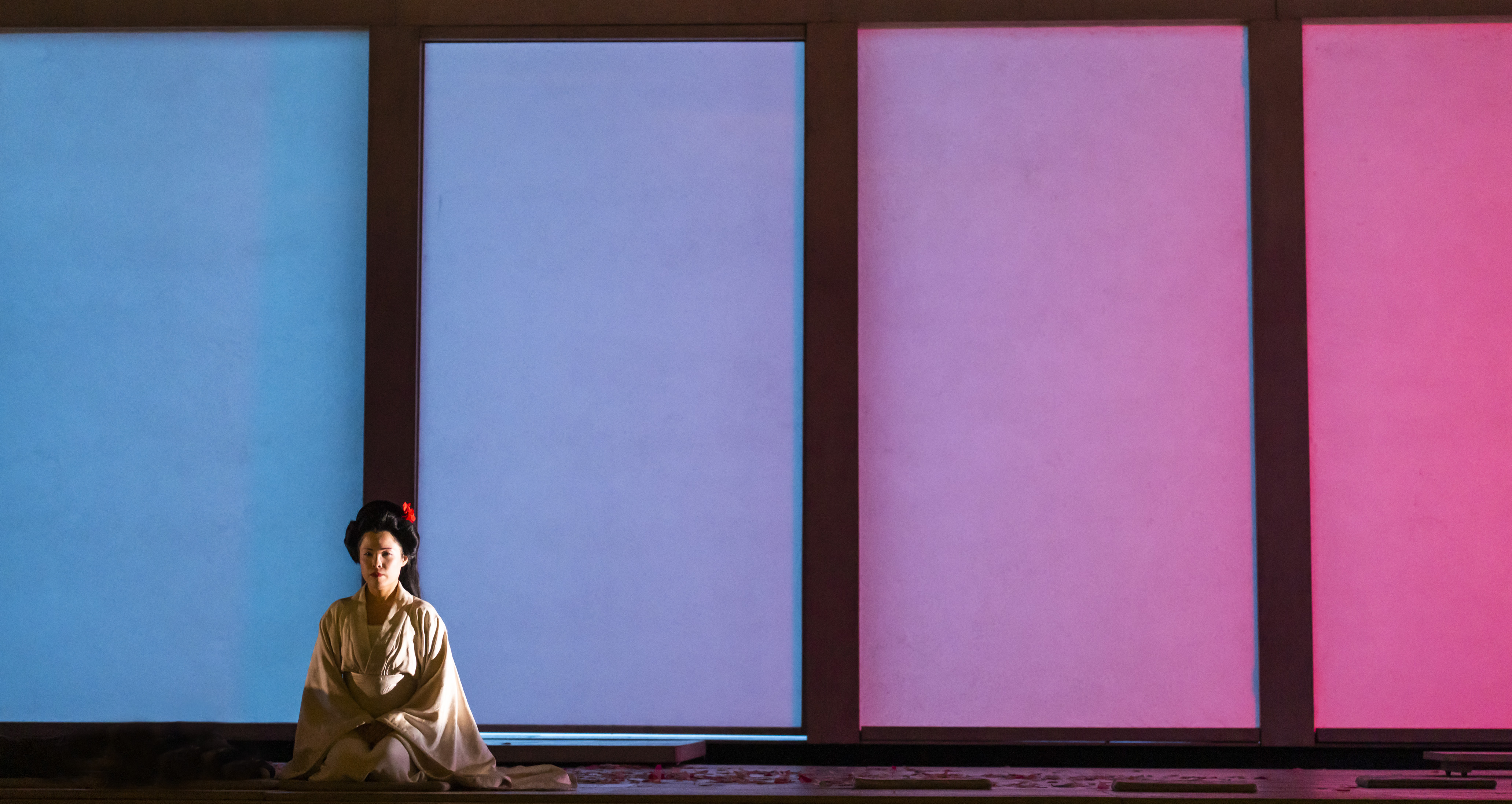 A performer in traditional Japanese Kimono sits in front of four windows that are lit with different colours. They are performing in The Royal Opera's production of Madama Butterfly. (4 of 6)