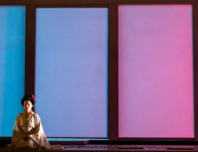 A performer in traditional Japanese Kimono sits in front of four windows that are lit with different colours. They are performing in The Royal Opera's production of Madama Butterfly. (4 of 6)