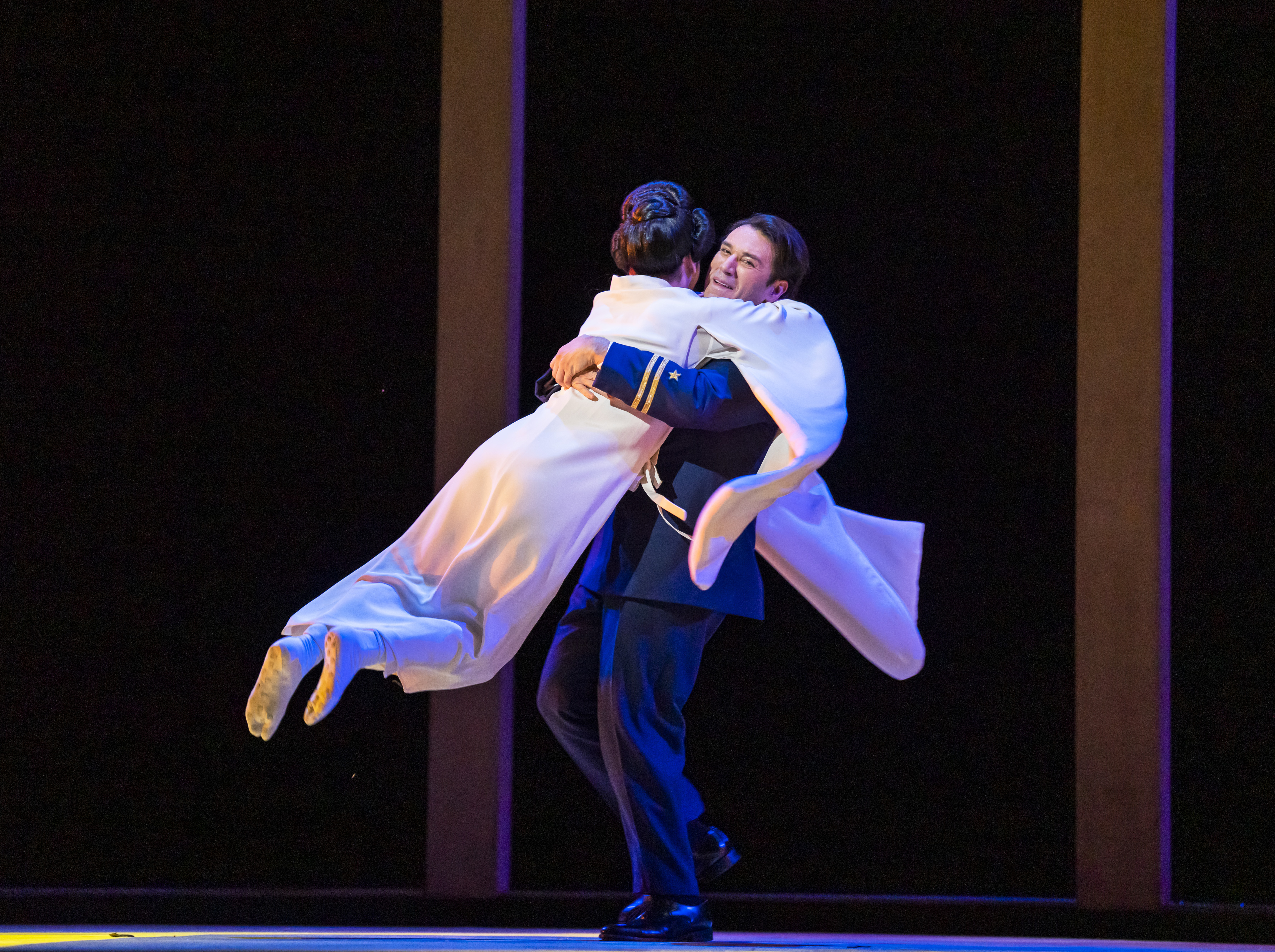A performer in a long white flowing Kimono jumps into the arms of a performer wearing a naval military uniform on stage. They are performing in The Royal Opera's production of Madama Butterfly. (3 of 6)