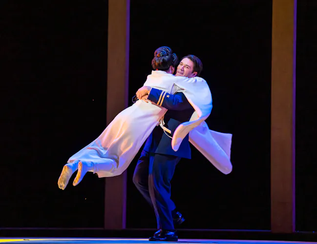 A performer in a long white flowing Kimono jumps into the arms of a performer wearing a naval military uniform on stage. They are performing in The Royal Opera's production of Madama Butterfly. (3 of 6)