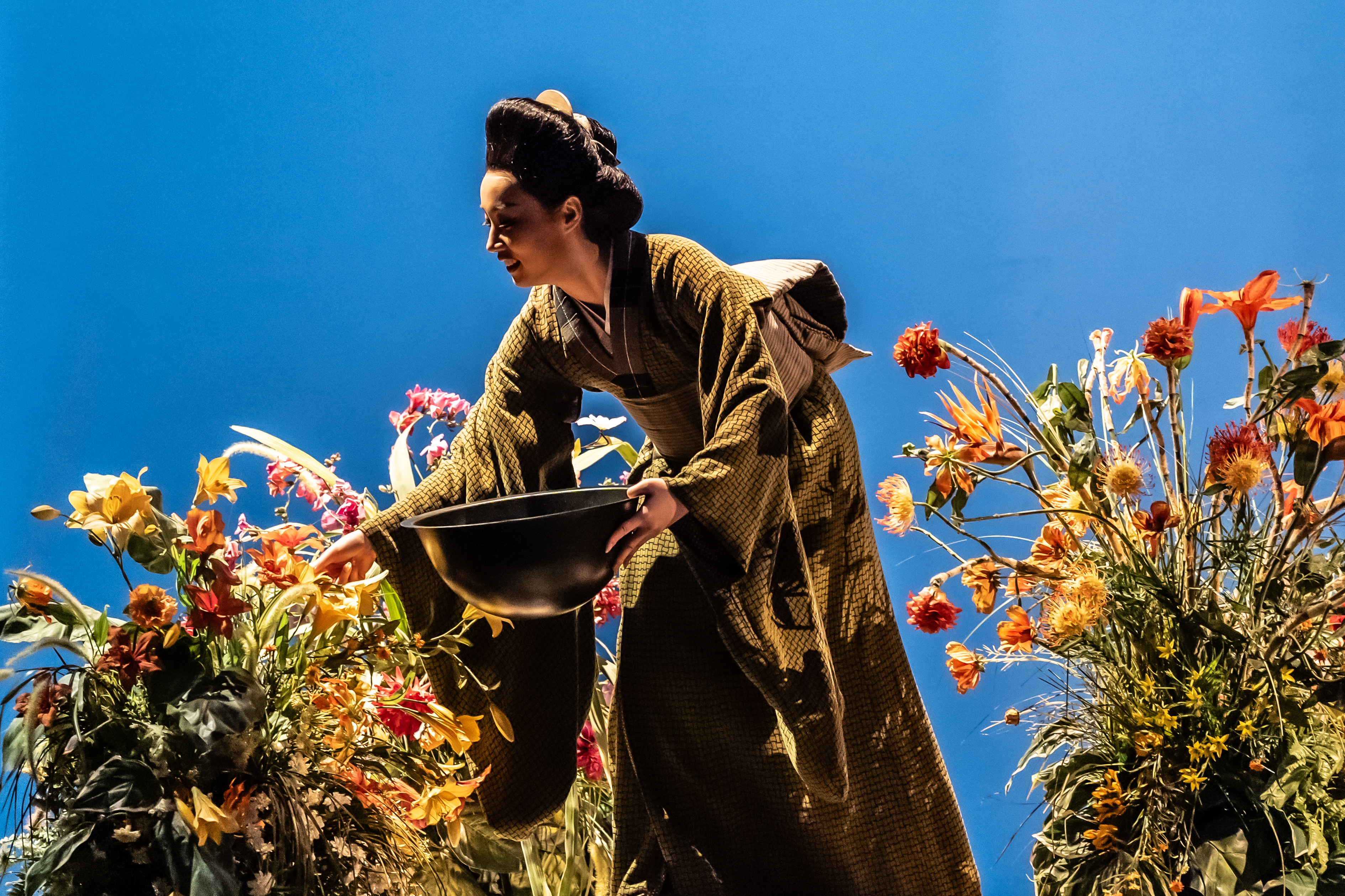 A performer wearing a Kimono holds a large bowl and picks from flowers scattered around a well-lit stage with a bright blue backdrop. They are performing in The Royal Opera's production of Madama Butterfly. (5 of 6)