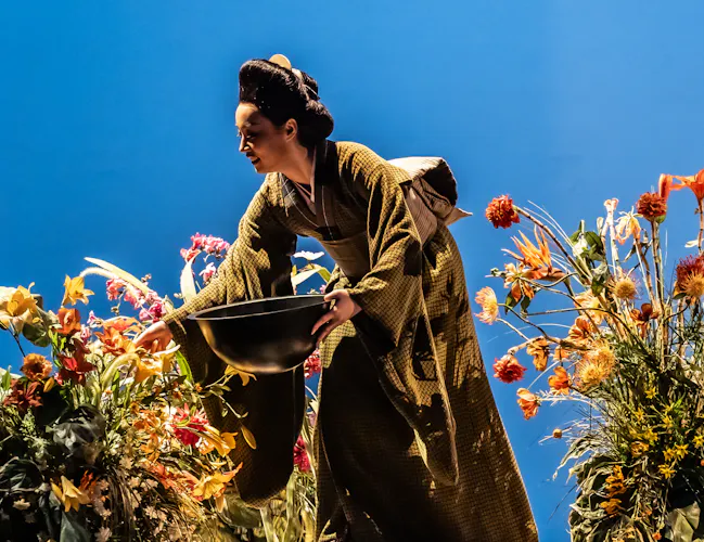 A performer wearing a Kimono holds a large bowl and picks from flowers scattered around a well-lit stage with a bright blue backdrop. They are performing in The Royal Opera's production of Madama Butterfly. (5 of 6)