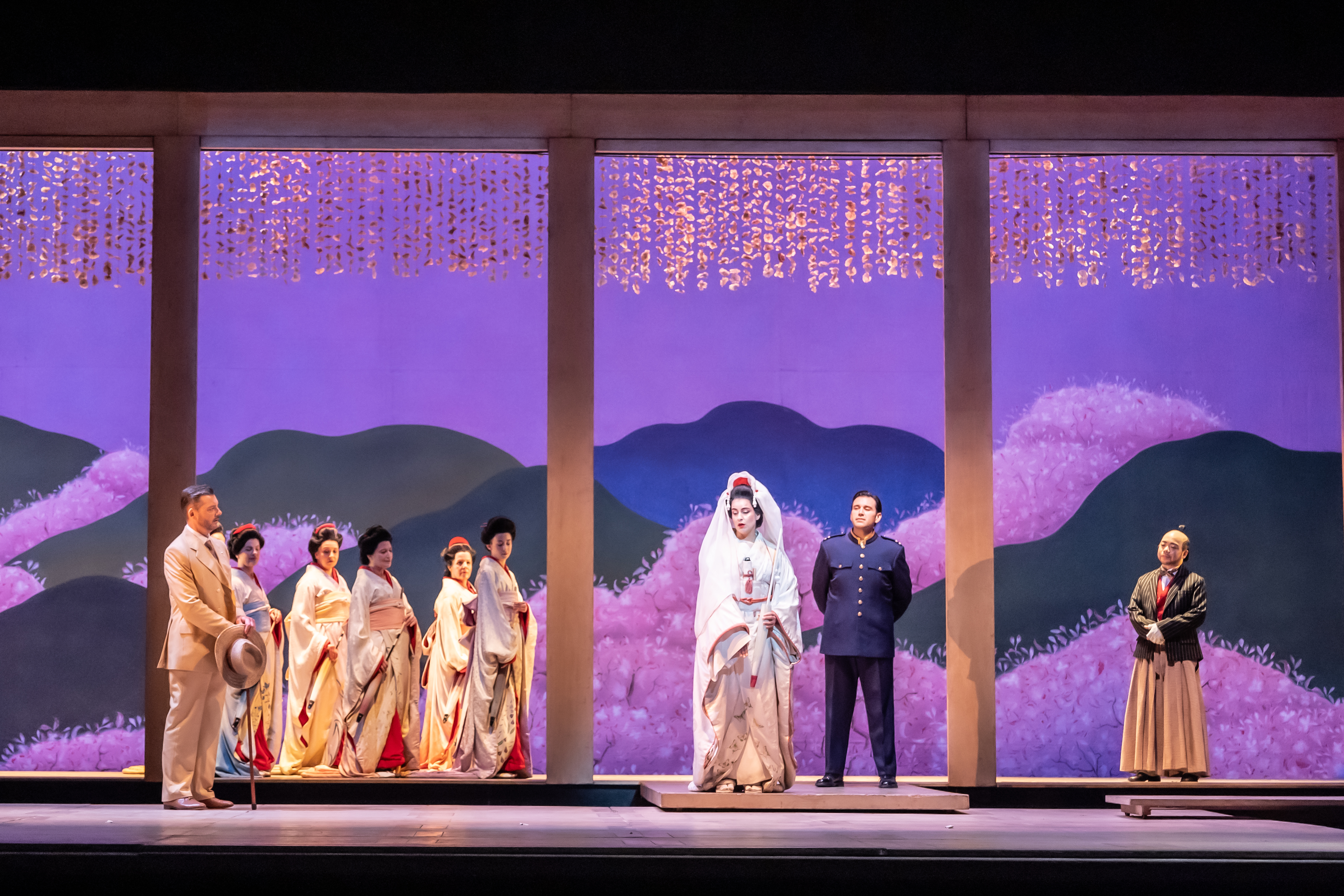 A group of performers stand on a brightly lit stage with a backdrop painted with purple mountains. The scene shows a wedding between two of the performers - one wears a white kimono and traditional headdress and the other wers a US Navy officer uniform. They are performing in The Royal Opera's production of Madama Butterfly. (6 of 6)