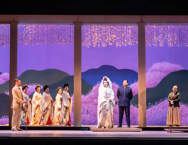 A group of performers stand on a brightly lit stage with a backdrop painted with purple mountains. The scene shows a wedding between two of the performers - one wears a white kimono and traditional headdress and the other wers a US Navy officer uniform. They are performing in The Royal Opera's production of Madama Butterfly. (6 of 6)