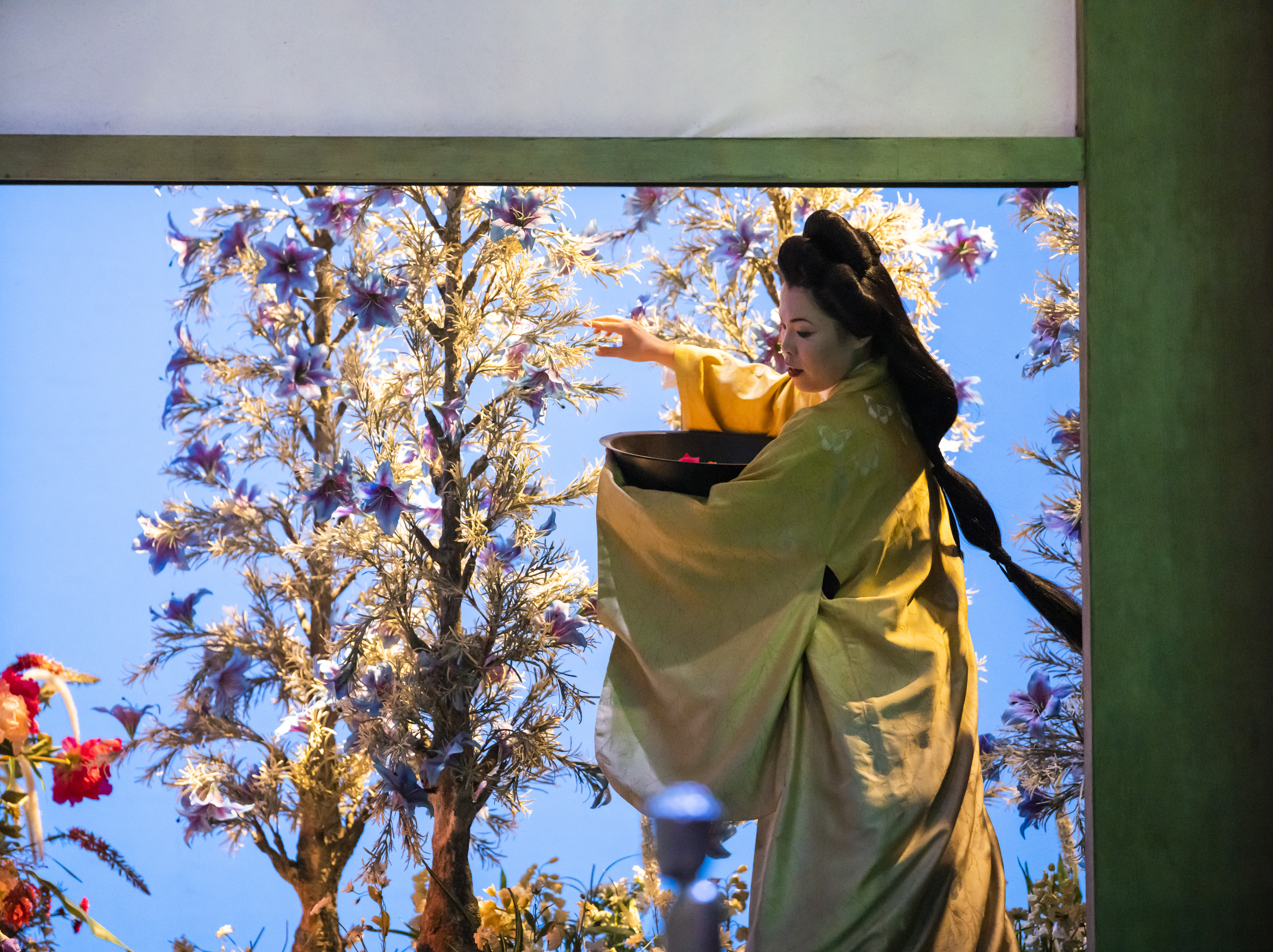 A performer wearing a kimono holds a basket in one arm and tends to tall flowers with the other on a well-lit stage with a bright blue backdrop. They are performing in The Royal Opera's production of Madama Butterfly. 